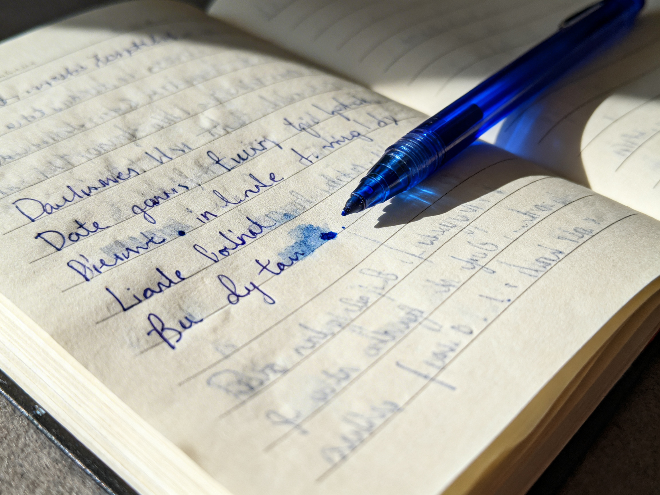 图中展示了一本摊开的米黄色横线笔记本，一支半透明的蓝色圆珠笔斜搁在页面上。阳光从侧面照射，在纸面上投射出笔的阴影，营造出一种午后书写的静谧氛围。笔记本上写满了蓝色的手写体文字，字迹略显潦草。然而，画面中存在明显的 AI 生成缺陷：首先，纸上的文字并非真实的语言，而是由扭曲、无意义的字母符号组成的“乱码”，且部分笔画与横线交织得极不自然；其次，笔尖下方有一块突兀的蓝色墨水渍，其晕染效果显得生硬且缺乏逻辑；最后，笔记本的横线在不同区域出现了透视断层和粗细不均的现象，远处的纸张边缘也显得模糊且结构模糊。整体构图虽然色彩清新，但细节处的逻辑错误揭示了其非实拍的本质。