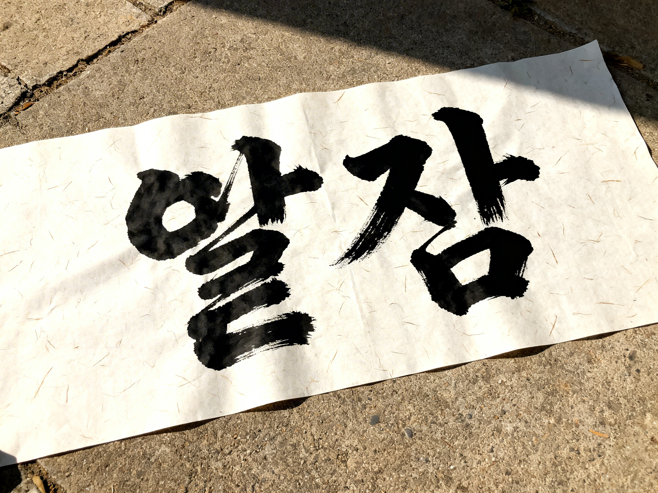 图中展示了一张铺在粗糙灰色水泥地面上的白色宣纸，纸上用黑色毛笔书写着两个巨大的韩文字符。阳光从右上方斜射而入，在纸张表面留下了清晰的阴影，纸张质感略显粗糙，隐约可见纤维纹理。构图采用俯视斜角，强调了文字的厚重感。然而，画面中存在明显的 AI 生成缺陷：首先，这两个韩文字符并非标准文字，其笔画结构混乱，像是将多种字符元素随意拼凑而成的“伪文字”，无法实际阅读。其次，左侧字符的笔触末端出现了不自然的断裂和重叠，墨迹的干湿浓淡变化不符合真实毛笔书写的物理逻辑。此外，纸张边缘与地面的交界处过于生硬，缺乏自然的厚度感，光影投射在褶皱上的表现也略显僵硬。