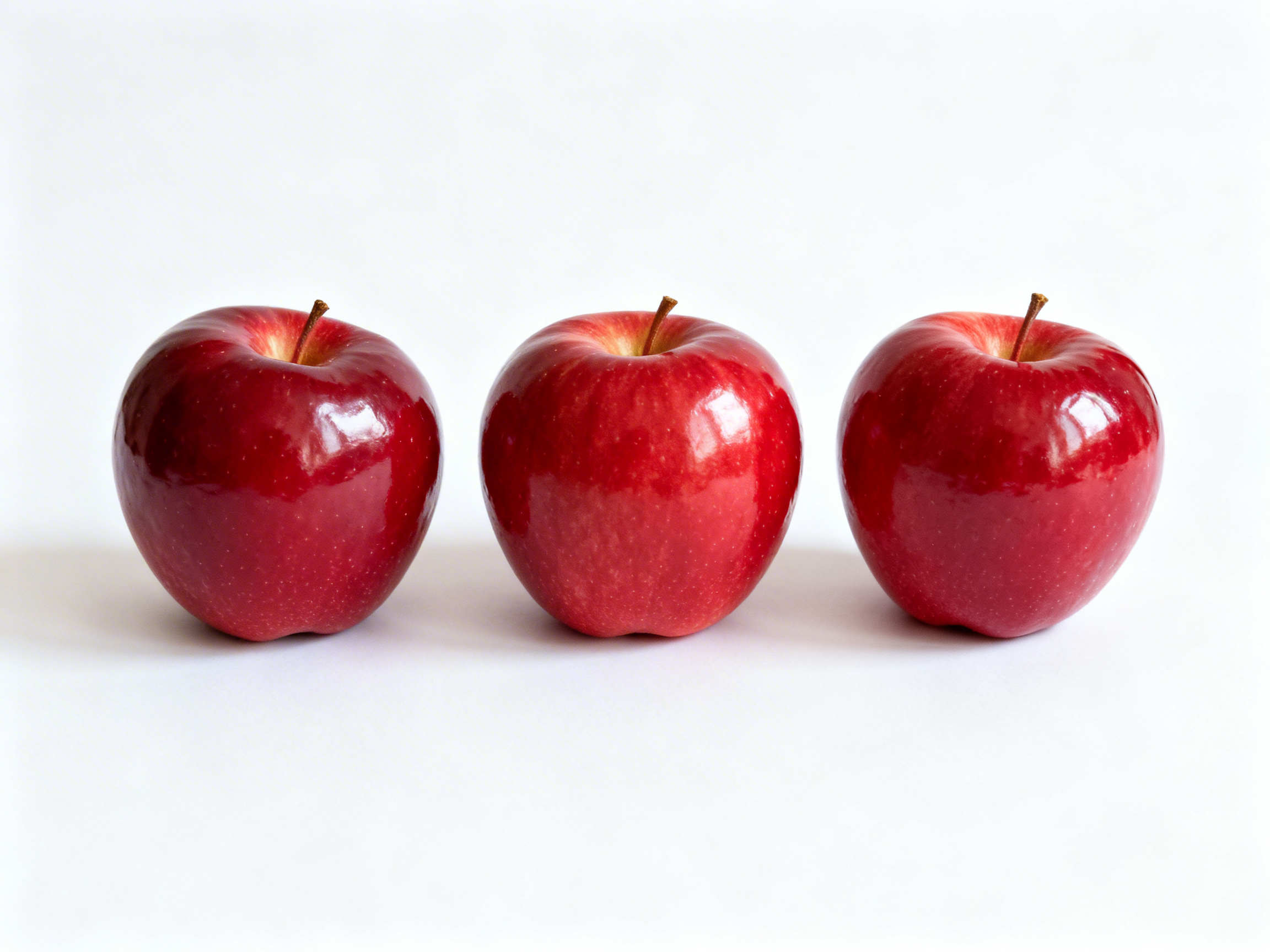 画面中，在纯白色的背景下，三颗鲜红色的苹果整齐地横向排列在画面中央。这些苹果色泽红润且富有光泽，表面带有细腻的浅色斑点，顶部都带有一根短小的褐色果梗。光线从右上方照射，使苹果表面出现了明显的白色反光，并在每个苹果的左下方投下了淡淡的柔和阴影。整体构图简洁对称，呈现出一种极简主义的静物美感。在 AI 生成的细节方面，虽然苹果的质感非常逼真，但仔细观察可以发现一些细微的逻辑异常：三颗苹果的形状和果梗的角度过于相似，缺乏自然界中果实应有的个体差异；此外，苹果底部与白色平面的接触点显得过于平滑，阴影的衔接略显生硬，缺乏真实的物理压痕感。