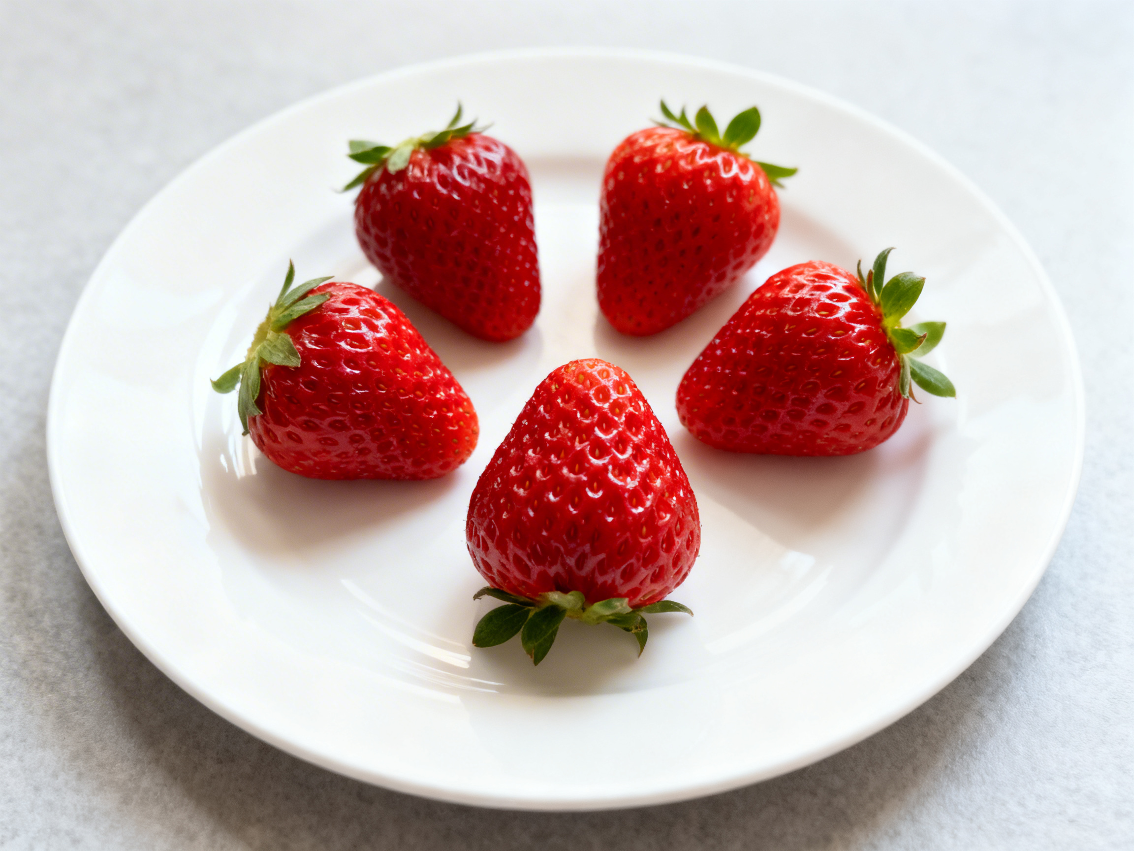 画面中展示了一个纯白色的圆形瓷盘，平放在浅灰色的纹理台面上。盘中整齐地摆放着五颗鲜红色的草莓，它们呈环形分布，色泽红润且表面带有自然的光泽感。每颗草莓顶部都保留着翠绿色的叶片。构图简洁，光线柔和，从左上方投射下淡淡的阴影。在 AI 生成的细节方面，存在一些明显的逻辑缺陷：位于画面最下方的一颗草莓，其绿色的叶片竟然生长在草莓尖端（底部），而非正常的顶端，这违背了植物的自然生长规律。此外，盘子右侧边缘的线条略显生硬，与背景台面的交界处透视感稍欠自然。整体而言，图片色彩明快，但在生物结构和空间细节上留下了 AI 生成的痕迹。