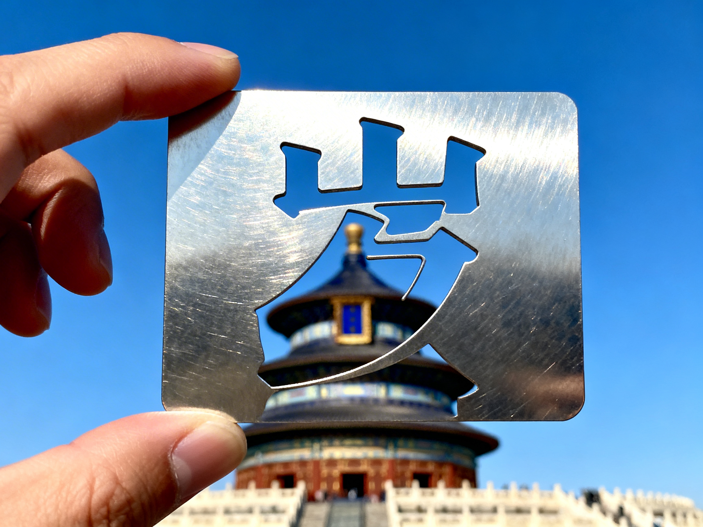 图中展示了一只手拿着一块长方形的金属镂空书签，背景是北京天坛的祈年殿。画面构图以手持书签为中心，书签表面有拉丝金属质感，中间镂空刻有一个汉字。透过镂空部分，可以清晰地看到远处蓝天下的天坛建筑。背景的天空呈明亮的湛蓝色，天坛建筑处于虚化状态，突出了前景的金属质感。

然而，这张 AI 生成的图片存在明显的逻辑和细节错误。首先，书签中心镂空的汉字形状异常，它看起来像是“岁”字的变形，但笔画结构完全错误，不符合任何标准汉字。其次，手部细节存在 AI 缺陷：左侧露出的手指关节连接生硬，指尖形状不自然；下方捏住书签的大拇指指甲盖边缘模糊，且与书签边缘的接触位置缺乏真实的物理受力感。此外，书签镂空边缘的透视与背景天坛的轮廓并未完美契合，呈现出一种生硬的拼贴感。