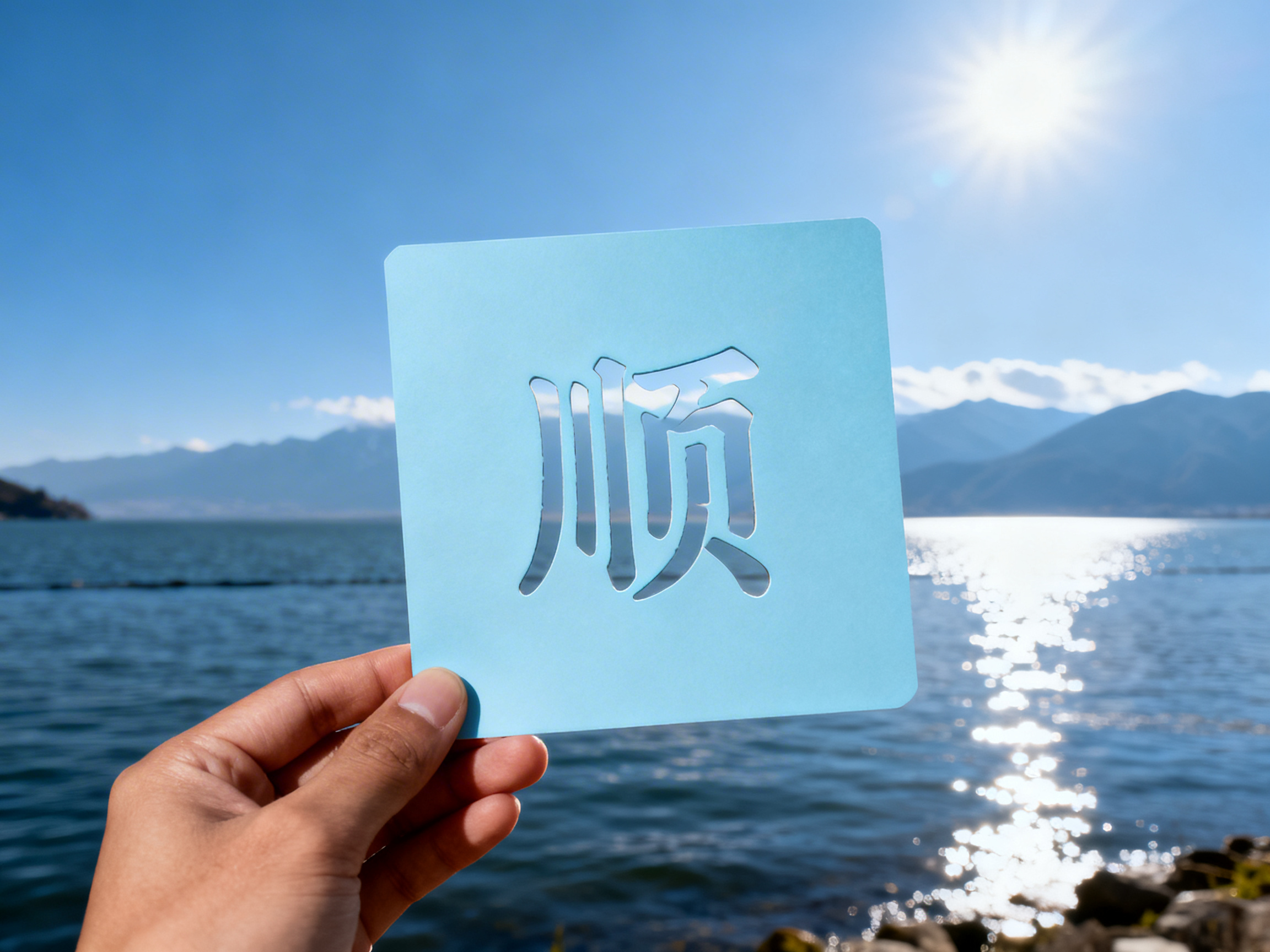 图中展示了一幅充满阳光的户外风景。画面前景中，一只左手捏着一张浅蓝色的正方形卡片，卡片中心镂空刻有一个汉字“顺”。透过镂空的字迹，可以看到背景中波光粼粼的水面。背景是一片开阔的湖泊或海面，远处有连绵的蓝色山脉，天空湛蓝清澈，右上角一轮烈日放射出耀眼的白光，光芒在水面上映照出细碎的金色亮斑。

这张图片具有明显的 AI 生成痕迹：首先是手部结构异常，拿卡片的手指比例不自然，大拇指与食指的连接处显得僵硬，且下方露出的其余手指数量和位置模糊不清，缺乏真实的骨骼感。其次，卡片上的“顺”字虽然清晰，但镂空边缘过于平滑且缺乏厚度感，与手部的光影衔接不够自然。此外，水面的波纹和远山的纹理略显重复，呈现出一种过度磨皮的数字平滑感。