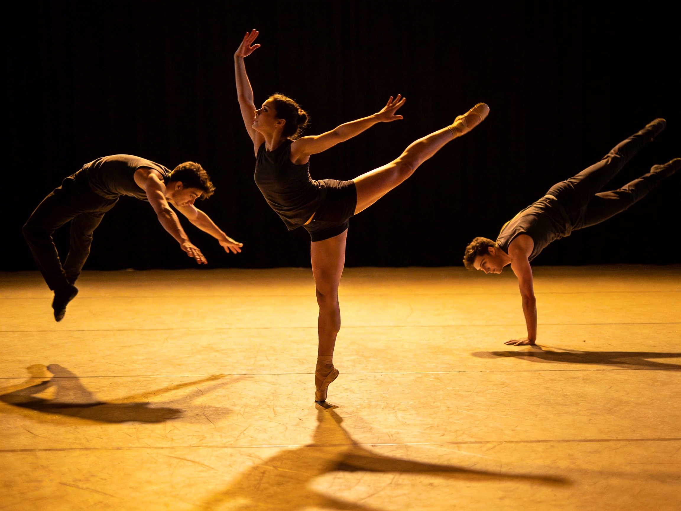 画面中展示了三位舞者在昏暗的舞台上表演现代芭蕾。背景是纯黑色的幕布，暖黄色的灯光从上方投射，在浅色的木质地板上拉出长长的影子。中心是一位女性舞者，她穿着黑色背心和短裤，单脚脚尖着地，另一条腿向后高高抬起，双臂舒展，姿态优美。她的左右两侧各有一名穿着黑色紧身衣的男舞者，左侧男子正向后跃起，右侧男子则单手撑地做出高难度的平衡动作。

然而，画面中存在明显的 AI 生成缺陷：最显著的是右侧男舞者，他仅靠一只右手支撑全身，但手臂线条与身体的连接显得生硬且不符合人体解剖学，且他的左臂完全缺失。中间女舞者的右手手指显得模糊且数量异常，左脚脚尖与地面的接触点透视关系略显生硬。左侧男舞者的左手手指也呈现出不自然的扭曲状。此外，地板上的影子与舞者的动作逻辑并不完全匹配，整体光影虽然氛围感强，但在细节处缺乏真实物理世界的严谨性。