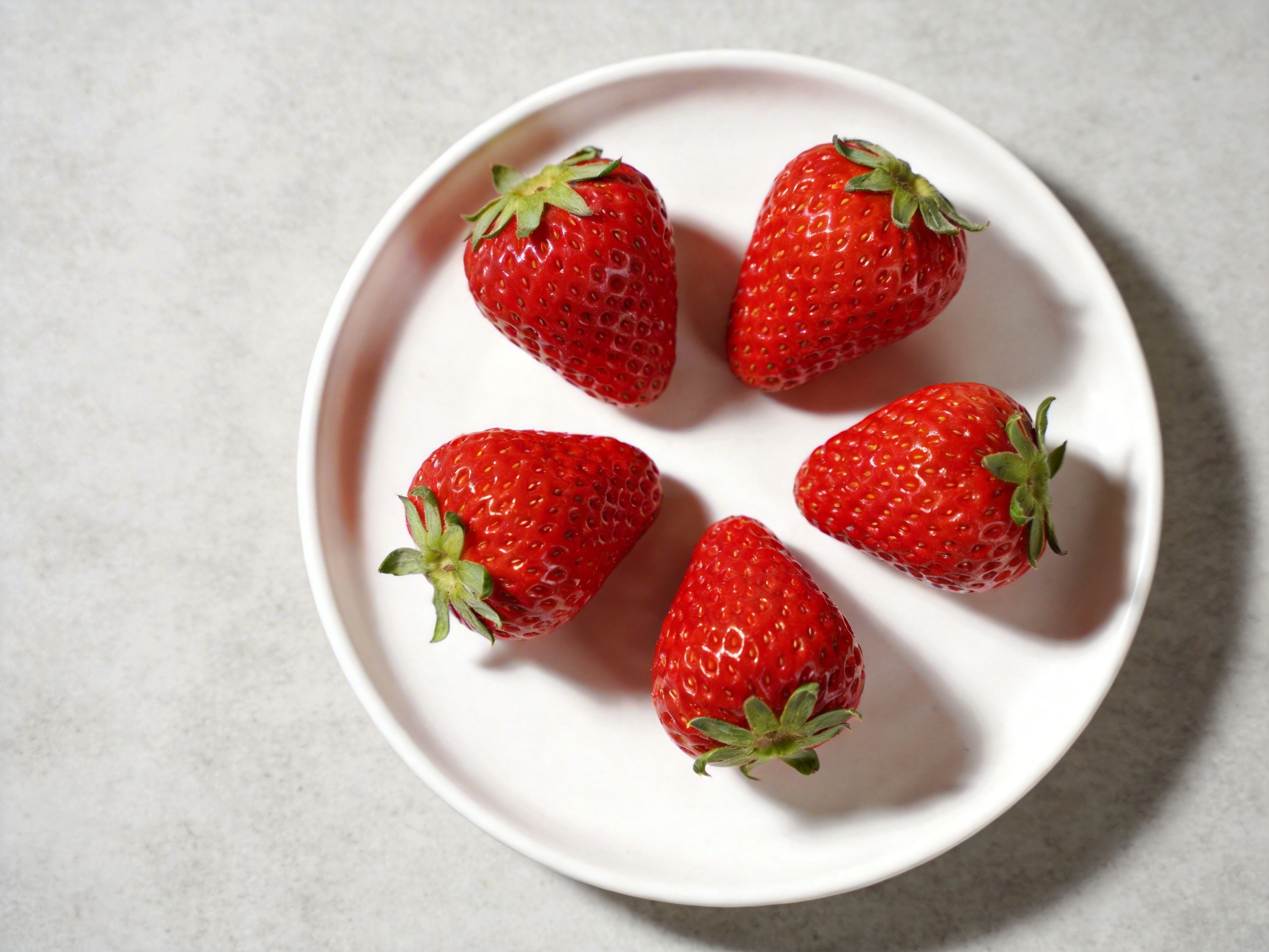 图中展示了一个简洁的俯视场景，在一个纯白色的圆形浅盘中，环状排列着五颗鲜红色的草莓。背景是带有细微颗粒感的浅灰色石质或水泥纹理桌面。草莓色泽红润饱满，表面布满了细小的黄色籽粒，顶端带有翠绿的叶片，光影效果显示光源来自左上方，使草莓在盘中投下了柔和的阴影。

虽然画面整体看起来非常写实，但仍能发现一些 AI 生成的典型缺陷：首先，草莓的形状过于规整且相似，缺乏自然生长中的个体差异；其次，最左侧草莓的绿色叶片与果实连接处显得有些生硬，缺乏自然的结构过渡；最明显的异常在于光影逻辑，盘子右侧边缘的阴影呈现出一种不自然的断层感，且五颗草莓的投影方向与盘子边缘的受光面并不完全吻合，暴露出光影合成的违和感。