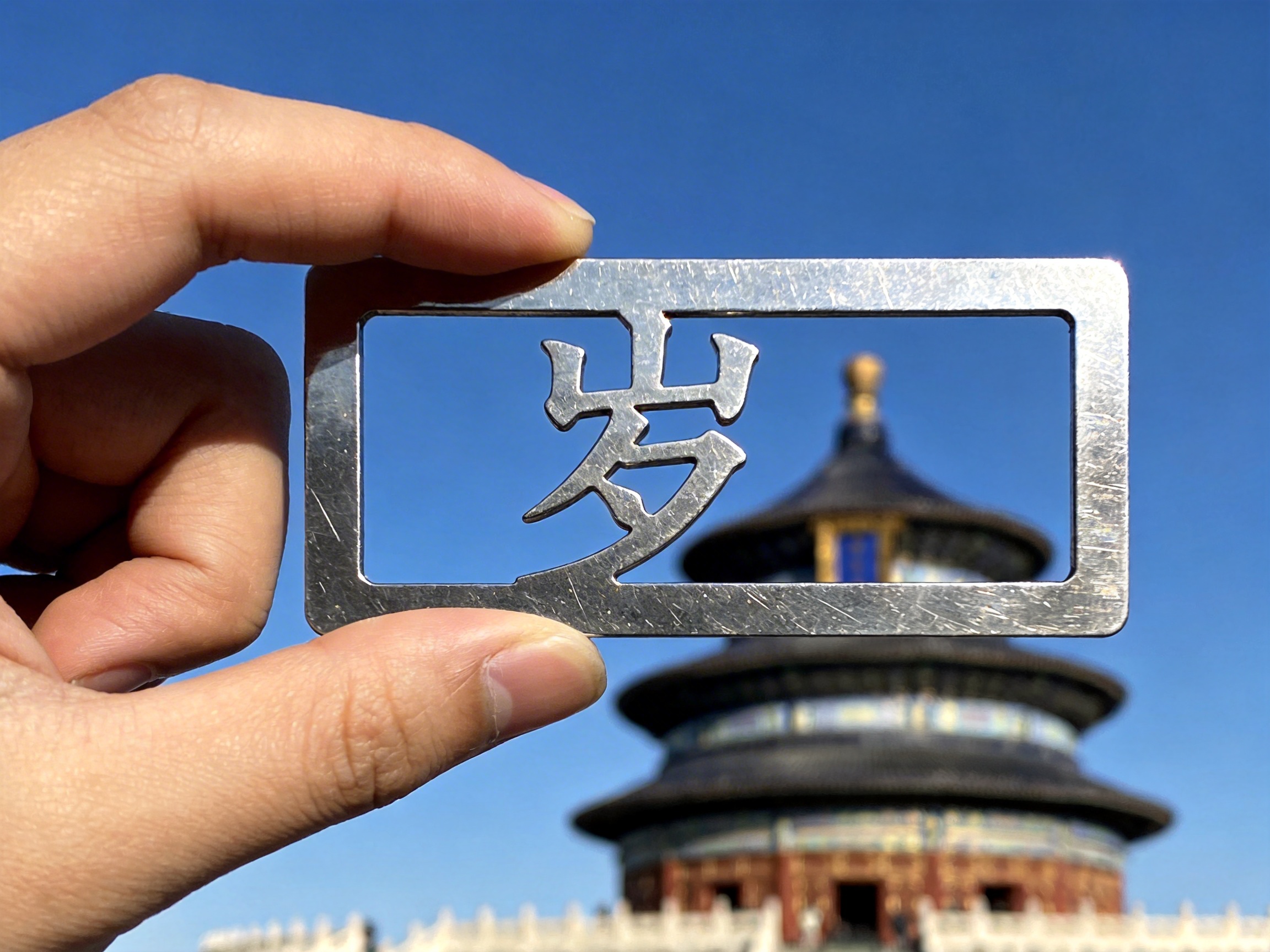 画面中，一只左手在湛蓝的晴空下举起一块长方形的金属镂空牌。金属牌表面带有细微的划痕质感，中心镂空出一个简体中文“岁”字。透过镂空的边框，背景中远处的北京天坛祈年殿被虚化处理，展现出蓝瓦金顶的宏伟轮廓。构图上，手部位于画面左侧，金属牌居中，与背景中的古建筑形成趣味对位。

然而，画面中存在明显的 AI 生成缺陷：首先，手部结构严重异常，在大拇指和食指捏住金属牌的同时，左侧边缘多出了一个无法解释的指节，且手指的生长方向和关节连接不符合人体解剖学。其次，金属牌中间的“岁”字笔画逻辑有误，上半部分的“山”字头与下半部分连接处显得生硬且不自然，缺乏真实的物理支撑感。此外，金属牌边缘的阴影与手指的接触面处理得较为模糊，透视关系略显违和。