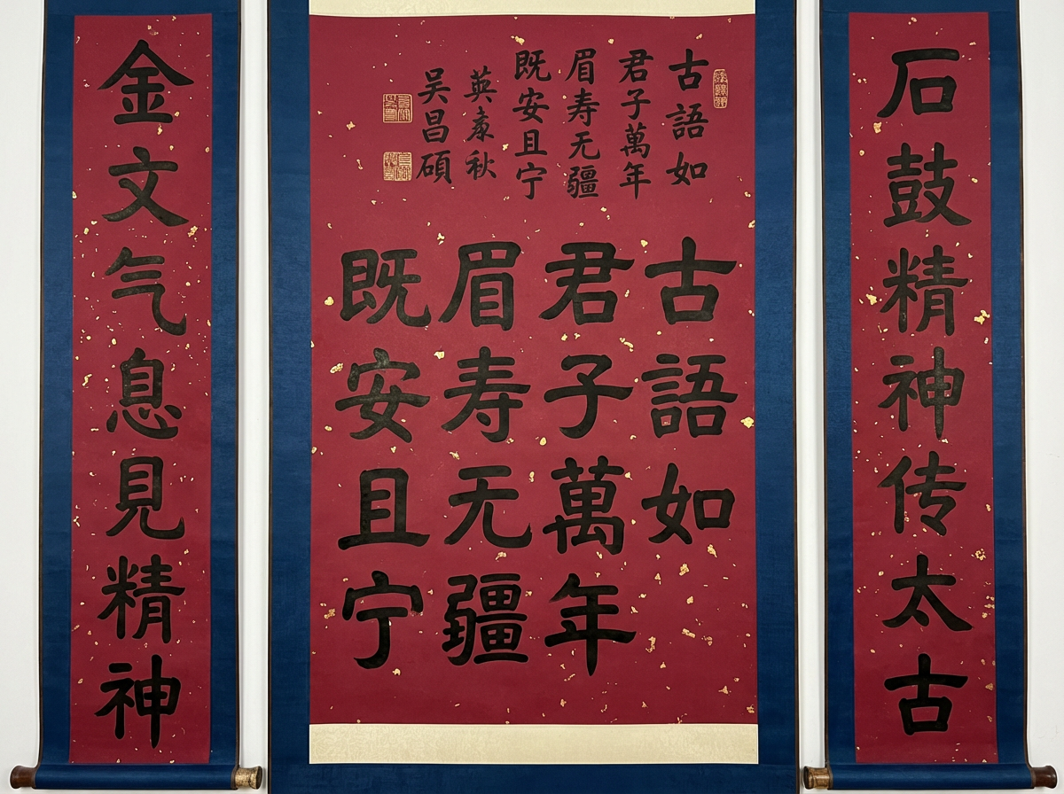 图中展示了一组由三幅卷轴组成的中国书法作品，悬挂在洁白的墙面上。卷轴采用了经典的红底金屑纸张，边缘镶嵌着深蓝色的绫布边框，整体色调庄重且富有节日气息。中间的主卷轴较大，书写着多行隶书字体，上方带有落款和红色印章；左右两侧则是对联形式的窄长卷轴。书法线条粗犷有力，墨色浓郁。

然而，画面中存在明显的 AI 生成缺陷。首先是文字逻辑混乱：左右对联的文字如“金文气息见精神”和“石鼓精神传太古”虽然字迹清晰，但中间主卷轴的文字出现了大量重复，且排版布局不符合传统书法的章法。其次，最显著的 AI 错误在于落款处的文字，出现了如“吴昌硕”等名字的扭曲变形，且印章的形状模糊、边缘不齐。此外，卷轴底部的木质轴头在透视和光影上显得不够自然，与墙面的衔接处略显生硬。