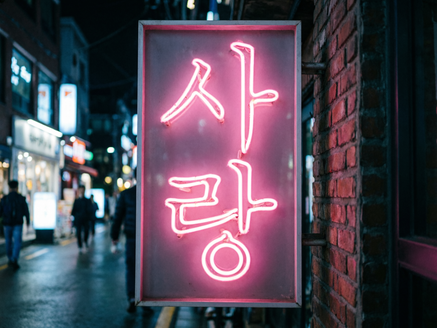 图中展示了一个夜晚的城市街道场景，焦点是一个悬挂在红砖墙上的长方形粉色霓虹灯牌。灯牌占据了画面的中心位置，上面闪烁着韩语词汇“사랑”（意为“爱”），粉红色的光芒在昏暗的背景中显得格外醒目。背景是虚化的街道夜景，可以看到模糊的行人背影和远处商店发出的冷色调灯光，地面似乎有些湿润，倒映着微弱的光。构图上采用了浅景深效果，使观众的注意力集中在灯牌的质感和色彩上。然而，作为一张 AI 生成的图片，画面存在明显的逻辑缺陷：灯牌上的韩文字形并不规范，笔画连接处显得生硬且不自然，尤其是下方的圆圈部分与上方笔画的衔接显得突兀。此外，灯牌左侧边缘的金属边框在透视上略显扭曲，与支撑架的连接方式也缺乏真实的物理结构感，背景中行人的轮廓过于模糊，甚至有些变形。