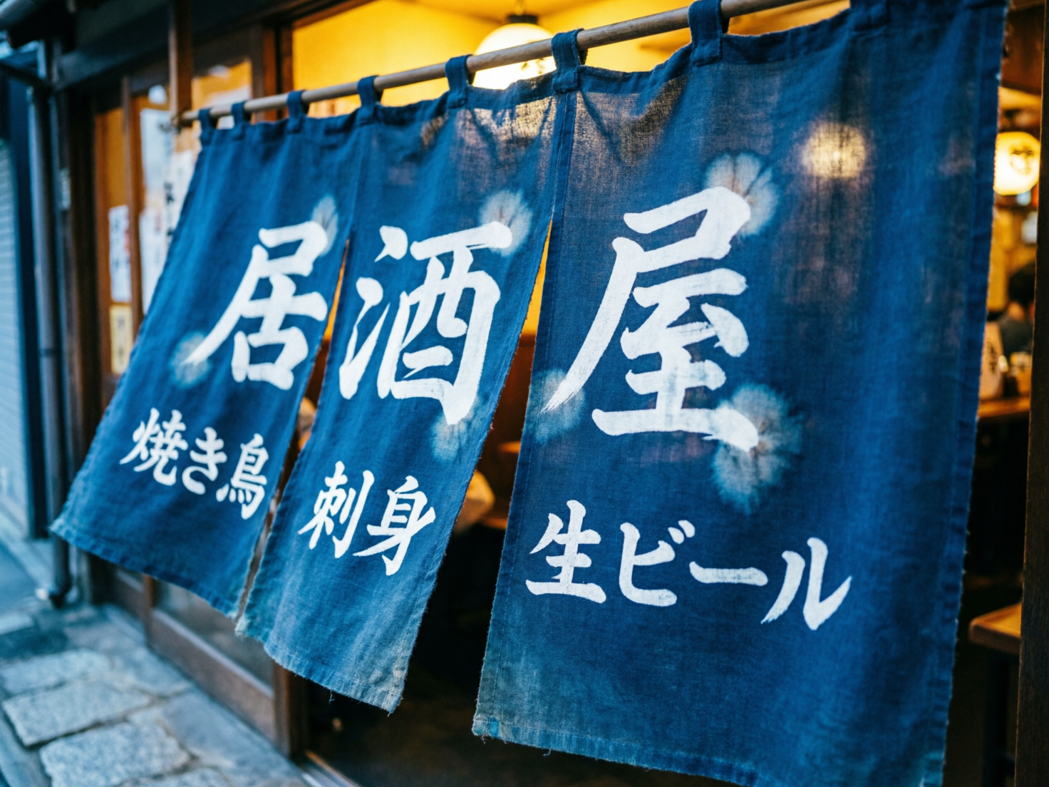 图中展示了一家日式居酒屋门前的近景。画面主体是挂在木质横杆上的三块深蓝色棉质暖帘，帘子上印有白色的手写体日文汉字。暖帘质感粗糙，带有扎染风格的白色圆点装饰。背景是居酒屋温暖的室内灯光，透出一种温馨的深夜食堂氛围。画面构图采用斜向透视，焦点集中在最右侧的帘子上。然而，这张 AI 生成的图片存在明显的文字逻辑缺陷：虽然大字写着“居酒屋”，但下方的细分菜单文字出现了混乱。最左侧本应是“烧鸟”的字样，其中“鸟”字的结构发生了扭曲变形；中间帘子上的“刺身”二字笔画生硬且不自然；最右侧的“生啤酒”日文片假名也存在笔画断裂和形态错误。此外，暖帘顶部的吊环与横杆的连接处显得有些模糊，缺乏真实的物理悬挂感。