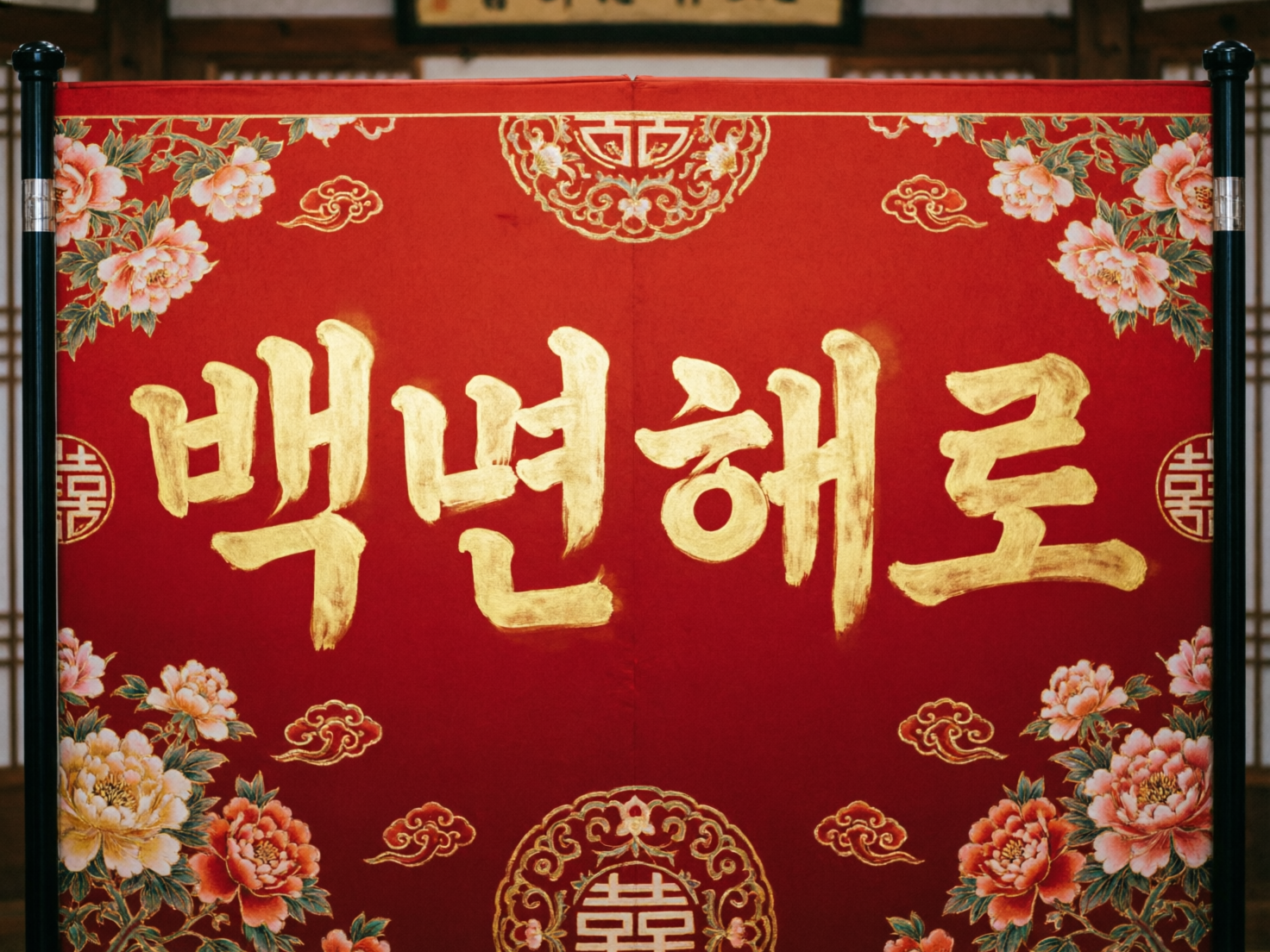 画面中展示了一块充满传统喜庆氛围的红色屏风或挂幅，背景似乎是一个古朴的室内空间。挂幅主体为鲜艳的大红色，正中心用金色书法字体书写了四个韩文字符“백년해로”，寓意“百年偕老”。挂幅的四个角落装饰着精致的粉色和橙色牡丹花纹，并点缀着金色的云纹和圆形的“囍”字图案。左右两侧各有一根黑色的金属支撑杆。

在 AI 生成的细节方面，存在一些明显的逻辑缺陷：首先，画面最下方的圆形“囍”字图案中，文字结构发生了扭曲和重叠，并非标准的汉字形态；其次，金色韩文书法的笔触边缘显得有些模糊，且与布料纹理的融合不够自然，带有明显的数字涂抹感；最后，挂幅顶部的边缘线条不够平直，左右两侧的支撑杆在透视和光影表现上略显僵硬，缺乏真实物体的质感。