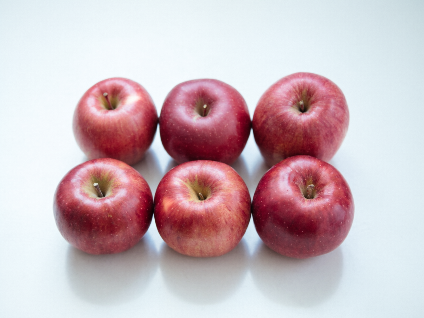 图中展示了六个红苹果，它们被整齐地排列成两行三列，摆放在一个纯净的白色平面上。这些苹果呈现出深浅不一的红色，表面带有细小的黄色斑点和自然的纹理，顶部的果梗凹陷处泛着淡淡的青黄色。光线从上方照射，在苹果下方的白色台面上投射出柔和的倒影和阴影，整体色调明亮且干净。

从 AI 生成的细节来看，画面存在一些不自然的缺陷：首先，苹果的果梗形态各异且显得生硬，部分果梗像是直接漂浮在凹陷处，缺乏真实的生长连接感。其次，苹果之间的透视关系略显违和，尤其是后排右侧的苹果与前排的间距在视觉上有些模糊。此外，苹果表面的反光分布过于均匀，缺乏真实物体在复杂光影下的质感变化，使得果皮看起来略显塑料感。