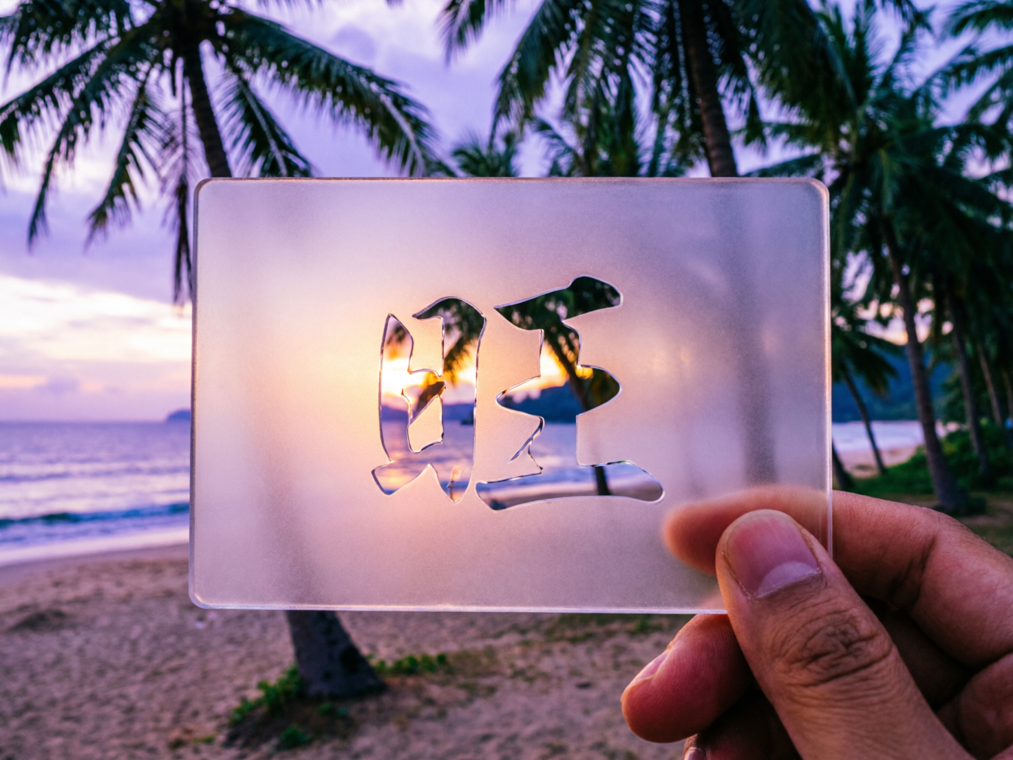 画面中展示了一张在海滩落日背景下拍摄的照片。一只肤色较深的手位于画面右下方，捏着一块半透明的磨砂质感方牌。方牌中心镂空刻有一个汉字，透过镂空处可以看到远处海平面上金灿灿的夕阳余晖。背景是热带海滨风光，沙滩宽阔，海浪拍打着岸边，上方点缀着几棵高大的椰子树，天空呈现出淡紫色与橙色交织的晚霞色彩。

画面中存在明显的 AI 生成缺陷：首先，方牌中心镂空的汉字结构极其混乱，看起来像是“日”字与“旺”字的错误结合，笔画断裂且不符合任何常规字体逻辑，属于典型的文字生成错误。其次，持牌的手部比例略显僵硬，指甲边缘与卡牌的接触面处理不够自然。此外，背景中左侧椰子树的叶片纹理显得过于细碎且分布杂乱，缺乏真实的物理生长结构。整体构图虽然意图营造唯美氛围，但由于核心元素的文字逻辑错误，显得不够真实。
