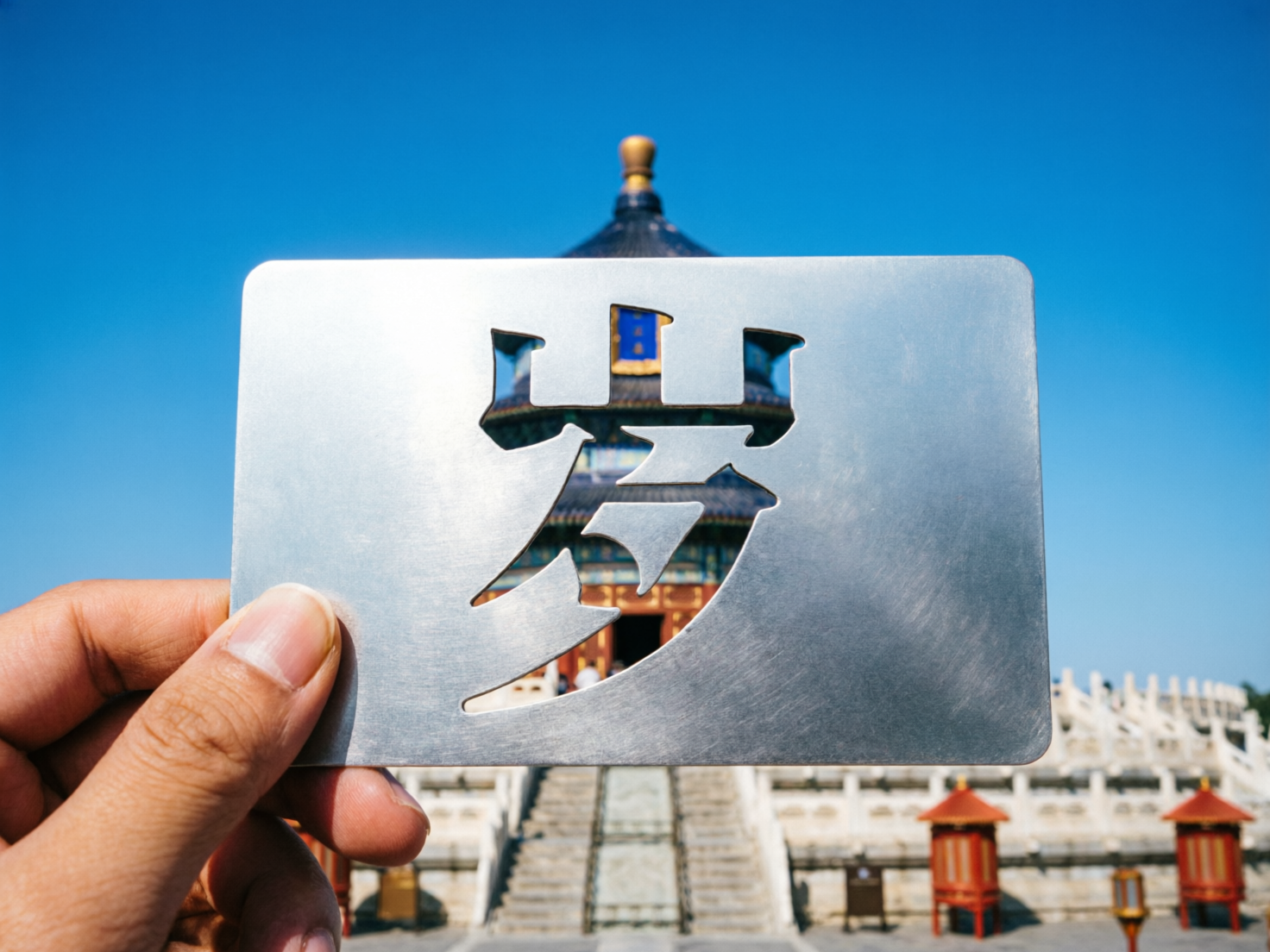 图中展示了一个晴朗蓝天下，一只手拿着银色金属镂空卡片对准北京天坛祈年殿的创意构图。卡片位于画面中央，镂空部分是一个巨大的汉字，但该字并非标准汉字，而是将「岁」字的下半部分与「万」字结合的错误字形，体现了 AI 在处理文字结构时的逻辑缺陷。透过镂空处，可以看到背景中被虚化的祈年殿，其蓝瓦红柱与金顶在阳光下格外醒目。左侧的一只手捏住卡片边缘，手指皮肤纹理清晰，但指甲形状略显生硬。背景的天坛建筑呈现出明显的透视异常，祈年殿的基座台阶与两侧的红色灯座在比例和空间衔接上显得不够自然，整体画面虽然色彩鲜艳、构思新颖，但在文字准确性和建筑透视上存在典型的 AI 生成痕迹。