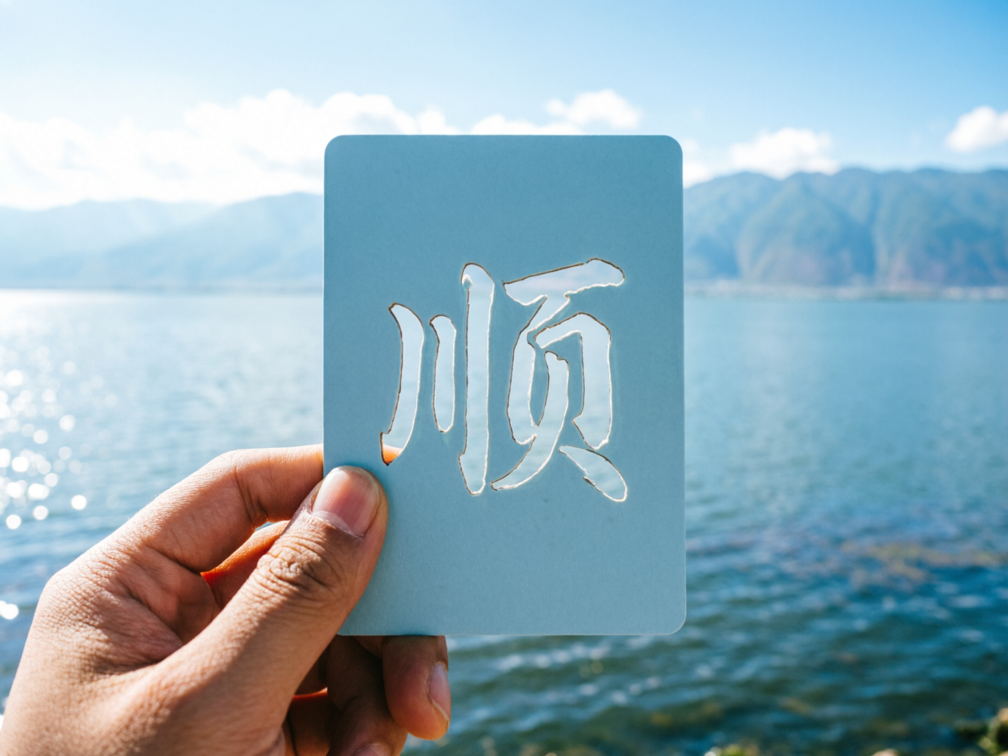 图中展现了一个充满阳光的户外场景。一只肤色偏深的手位于画面左下方，正捏着一张淡蓝色的长方形卡片。卡片的中心被镂空刻出了一个汉字“顺”，透过镂空的笔画可以看到背景中波光粼粼的湖水。背景由近及远分别是碧蓝的湖泊、连绵起伏的青色山脉以及晴朗的天空，整体色调清新自然，给人以宁静致远的感觉。

在 AI 生成的细节方面，存在一些明显的逻辑缺陷：首先，镂空的汉字“顺”在物理结构上是不合理的，例如右侧“页”字中间的封闭区域在现实中应该会脱落，但图中却悬浮在空中，没有连接点。其次，持卡的手部细节略显生硬，大拇指的指甲形状与关节比例不够自然。此外，卡片左侧边缘与手指接触的地方，透视关系略显模糊，卡片像是直接贴在手上而非被真实捏住。