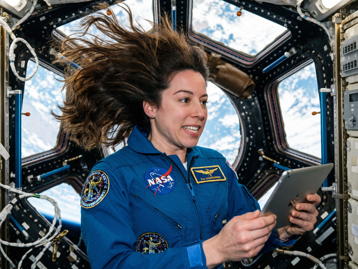 图中展示了一位身穿蓝色 NASA 宇航服的女性宇航员，正身处国际空间站的穹顶舱内。她拥有棕色长发，头发在失重状态下向斜上方飘散，神情显得惊喜且专注，手中正拿着一台平板电脑。背景是空间站标志性的多窗结构，透过窗户可以看到蔚蓝的地球弧线和白云。画面构图紧凑，光线明亮，呈现出一种科幻而真实的氛围。

然而，画面中存在明显的 AI 生成缺陷：最显著的是宇航员的双手，手指数量异常且关节扭曲，尤其是握持平板电脑的左手，手指显得杂乱且不符合人体解剖结构。此外，宇航服上的 NASA 标志和徽章虽然色彩鲜艳，但其中的文字和图案细节模糊、杂乱，并非真实的文字。空间站内部的电缆和机械结构在边缘处也存在透视逻辑错误，部分线条凭空消失或衔接不自然。