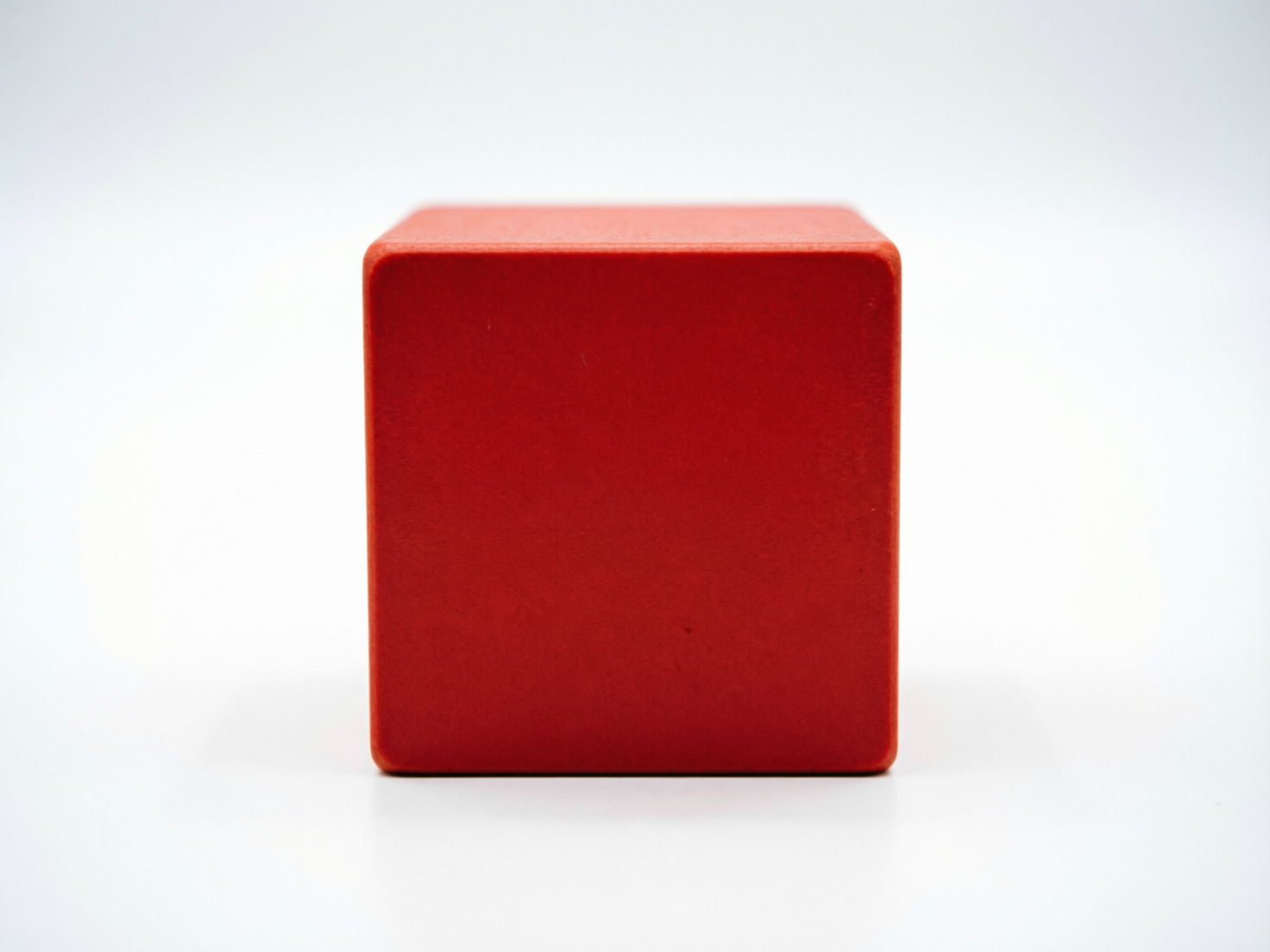 图中展示了一个位于画面中心的正方体木块，背景为纯净的浅灰色至白色渐变。木块呈现出鲜艳且饱和的红色，表面具有细微的磨砂质感，边缘处理得较为圆润。构图非常简洁，采用正面平视的角度，木块占据了画面的核心位置，底部带有淡淡的投影，营造出一种悬浮或放置在平面上的立体感。在 AI 生成的细节方面，虽然整体造型规整，但仔细观察可以发现，木块顶部的透视线条与正面的垂直边缘在衔接处略显生硬，顶面的纹理分布不够自然，且左下角的边缘轮廓线与背景交界处存在轻微的锯齿感，这些细微的几何偏差揭示了其 AI 生成的痕迹。总体而言，这是一张极简主义风格的单体物件展示图。