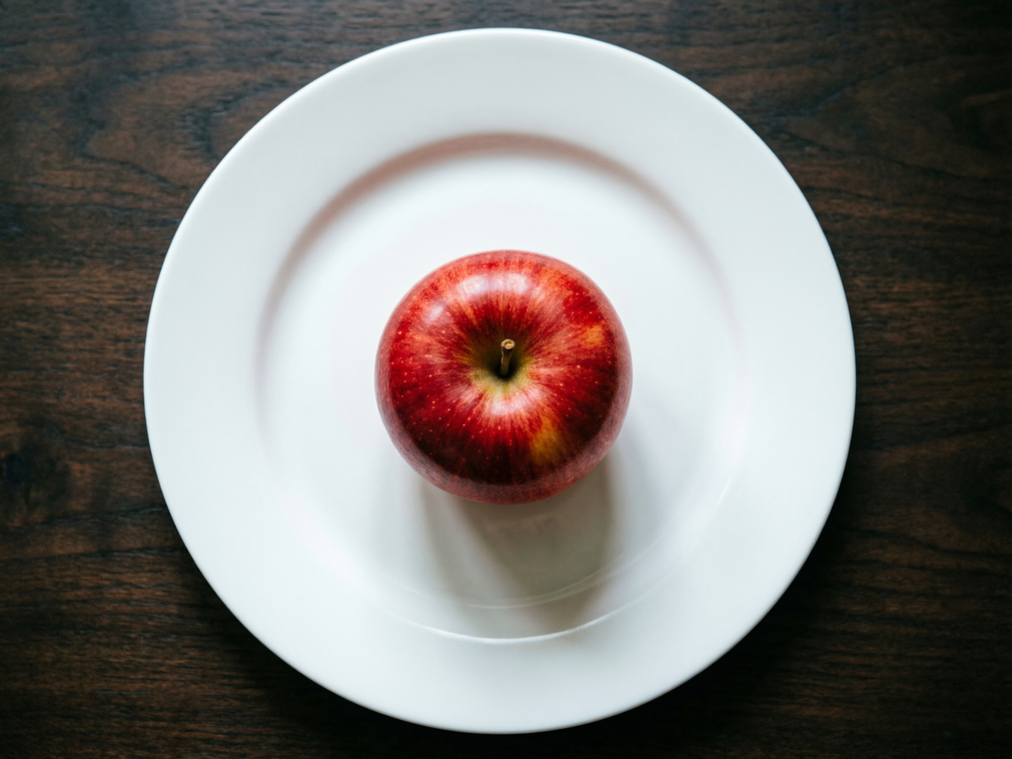 图中展示了一个极简主义风格的静物场景。画面正中央是一个圆形的白色陶瓷平盘，盘子中心放置着一颗饱满的红苹果。背景是深棕色的木质桌面，木纹清晰可见，与洁白的盘子形成鲜明对比。构图采用了垂直俯视的视角，呈现出一种对称的几何美感。苹果表面带有自然的红色条纹和细微的黄色斑点，顶部的果梗清晰可见，光线从上方照射，在苹果下方投射出柔和的阴影。

作为一张 AI 生成的图片，画面整体表现得非常逼真，但在细节处仍存在一些透视异常。仔细观察可以发现，盘子边缘的厚度在不同角度显得略有不均，且盘子中心与苹果接触位置的阴影处理显得有些生硬，缺乏真实物理环境下的自然过渡。此外，苹果顶部的凹陷处与果梗的连接方式略显僵硬，不够自然。尽管如此，整幅画面色彩明快，视觉重心非常突出。