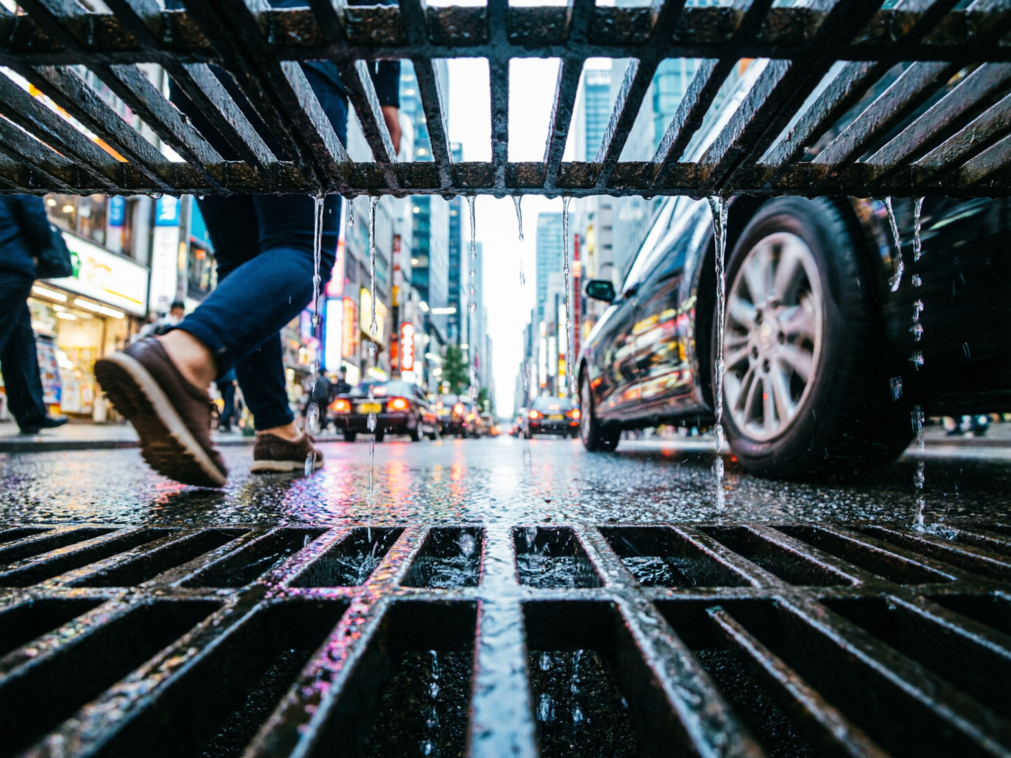 图中展示了一个从城市排水沟内部向上仰视的独特视角。画面构图被上下两层黑色的金属格栅切分，水滴正从上层格栅滴落。背景是繁华的都市街道，左侧有一名穿着深色长裤和棕色皮鞋的行人正迈步走过，右侧停靠着一辆黑色轿车，远处可见密集的建筑和闪烁的霓虹灯招牌。地面湿漉漉的，倒映着城市的灯光，整体色调偏冷并带有鲜艳的灯光点缀。

画面中存在明显的 AI 生成缺陷：首先，透视逻辑存在严重错误，观察者仿佛身处排水沟“内部”，但上下两层格栅的间距和位置在现实物理空间中难以成立；其次，左侧行人的腿部与身体连接处显得僵硬且比例失调，鞋底的纹路模糊不清；右侧汽车的车轮毂细节混乱，线条扭曲；最显著的是水滴的形态，它们像透明的柱状物一样垂直悬挂在空中，缺乏真实的重力感和动态水花的逻辑。