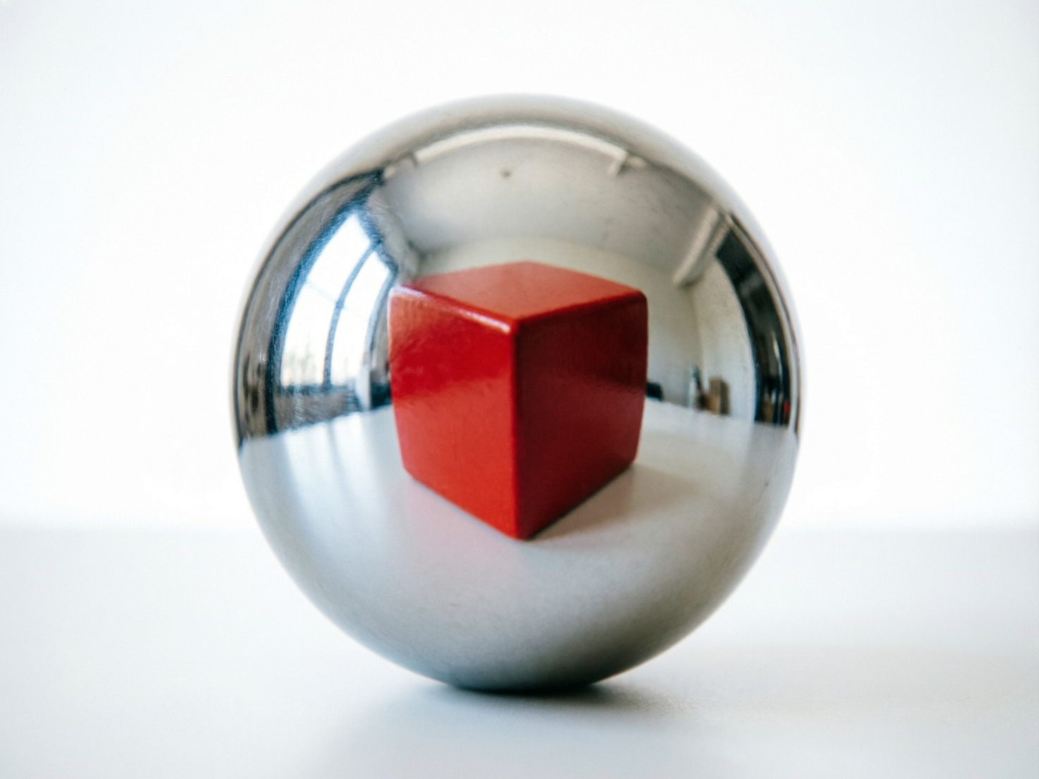 图中展示了一个极简主义风格的构图，主体是一个放置在白色平面上的巨大金属抛光球体。球体表面具有极高的反射率，呈现出冷色调的银灰色，并清晰地映射出周围室内环境的倒影，包括窗户的亮光和模糊的家具轮廓。最引人注目的是，球体中心反射出一个鲜红色的正方体，其色彩饱和度极高，与银色的球体形成鲜明对比。构图采用中心对称，背景是纯净的白色，营造出一种超现实且冷峻的艺术氛围。在 AI 生成的细节方面，存在明显的逻辑缺陷：球体反射出的红色立方体在现实空间中并不存在（球体前方是空的），这种“凭空出现”的反射违背了物理光学常识。此外，球体底部的接触面阴影处理略显生硬，缺乏真实的重力感。