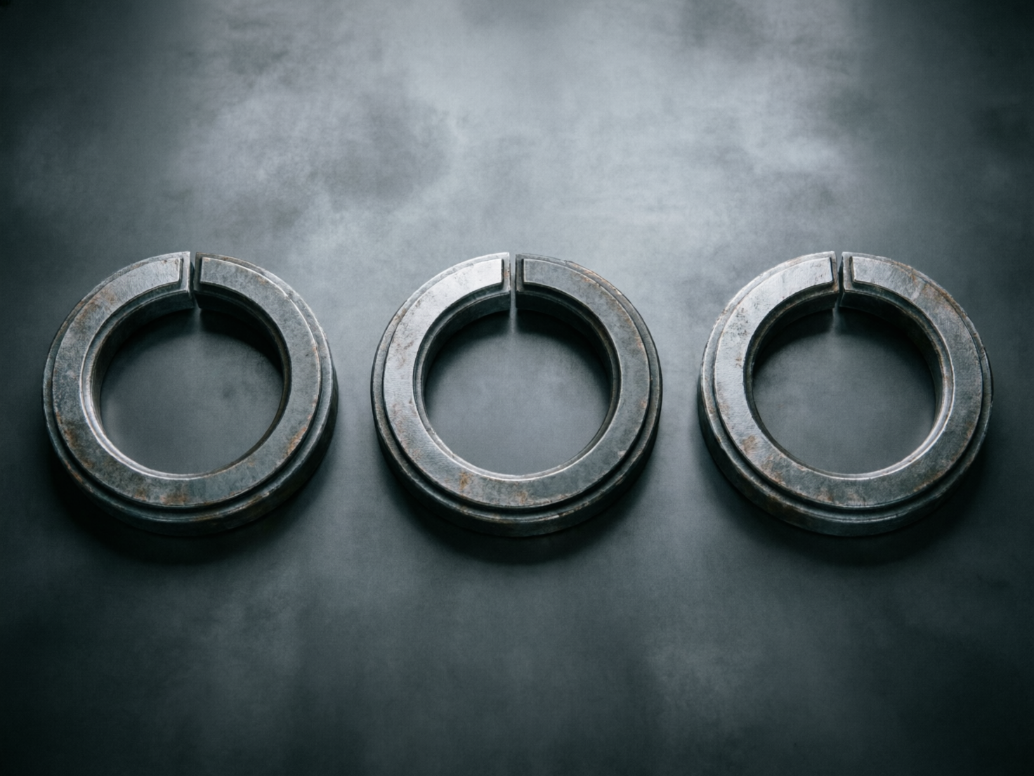 画面中展示了三个并排水平排列的厚重金属圆环，放置在一个带有磨砂质感的深灰色平面上。这些圆环呈现出冷峻的工业风格，表面有明显的磨损、锈迹和金属拉丝纹理，色调以深灰和银色为主。每个圆环的顶部都有一个垂直的切口，使其看起来像是一个未闭合的开口环或某种机械垫圈。光线从上方照射，在圆环下方投射出柔和的阴影，增强了物体的立体感。

作为一张 AI 生成的图片，画面中存在一些逻辑和透视上的瑕疵：首先，三个圆环顶部的切口形状并不完全一致，中间圆环的切口显得过于规整且缝隙较窄，而左右两侧的切口边缘略显模糊。其次，圆环表面的锈斑分布显得有些刻意和重复。最明显的缺陷在于透视关系，虽然三个圆环处于同一平面，但最右侧圆环的厚度与左侧两个相比略显单薄，且其内圈的阴影处理与整体光源方向不完全吻合，显得不够自然。