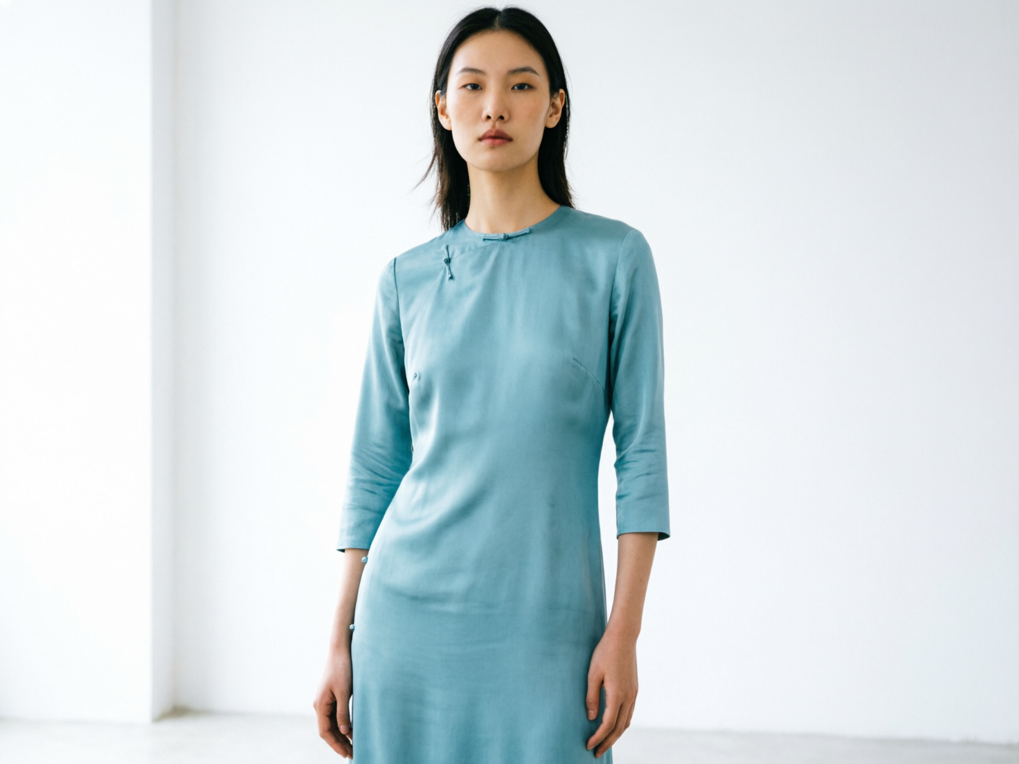 图中展示了一位具有东亚面孔特征的年轻女性，她正面对镜头站立在纯白色的简约室内空间中。她留着一头黑色中长发，表情冷峻且端庄。她穿着一件浅蓝色的丝绸质地长裙，裙子融合了中式旗袍的设计元素，领口处有精致的盘扣。光线从侧方柔和地打在她的身上，营造出一种极简主义的时尚感。

画面中存在明显的 AI 生成缺陷：首先，人物的手部结构异常，尤其是画面左侧（人物的右手）手指线条模糊且融合在一起，缺乏清晰的指节；其次，衣服的剪裁和褶皱在胸部和肩膀处显得不够自然，左肩处的缝合线略显生硬。此外，人物的双眼眼神略显空洞，瞳孔的对称性存在细微的偏差，背景墙壁与地面的交界处也显得过于平滑，缺乏真实环境的质感。