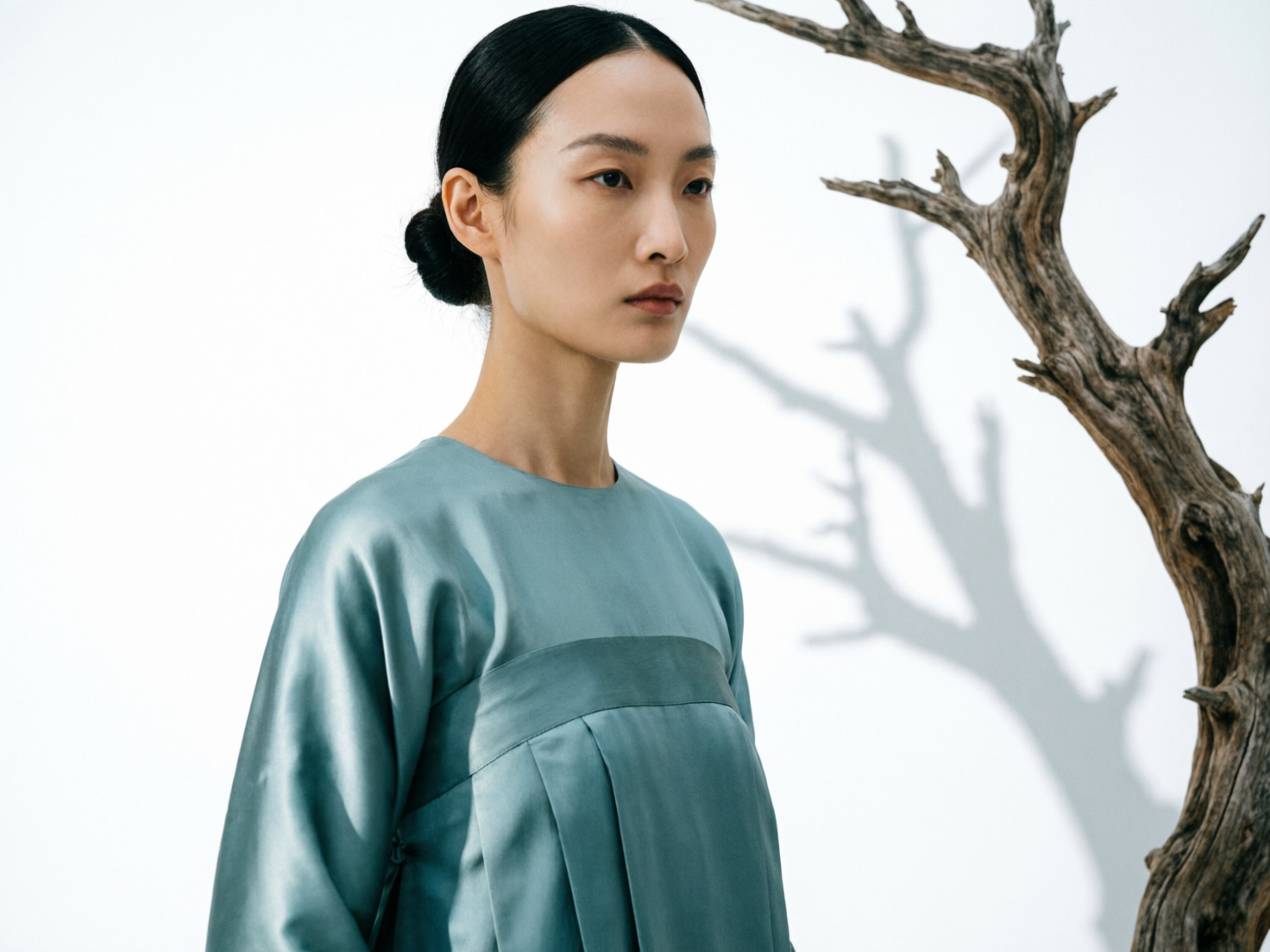 图中展示了一位具有东方韵味的女性模特，她侧身站立在纯白色的背景前。模特留着整洁的中分黑发，并在脑后盘成发髻，面部轮廓清冷，眼神忧郁地望向画面右侧。她穿着一件质地丝滑、带有光泽的青蓝色绸缎长袍，胸前有褶皱设计，整体风格简约而典雅。画面右侧矗立着一株枯木，其苍劲的枝干向斜上方延伸，并在白色背景上投下了清晰的树影，营造出一种禅意与孤寂的氛围。在 AI 生成的细节方面，存在一些明显的逻辑缺陷：模特左侧肩膀处的衣袖剪裁显得生硬且不符合人体结构，呈现出一种扁平的纸片感；此外，背景中树枝的投影与实物的形状并不完全匹配，阴影的透视关系略显杂乱，缺乏真实自然的光影逻辑。