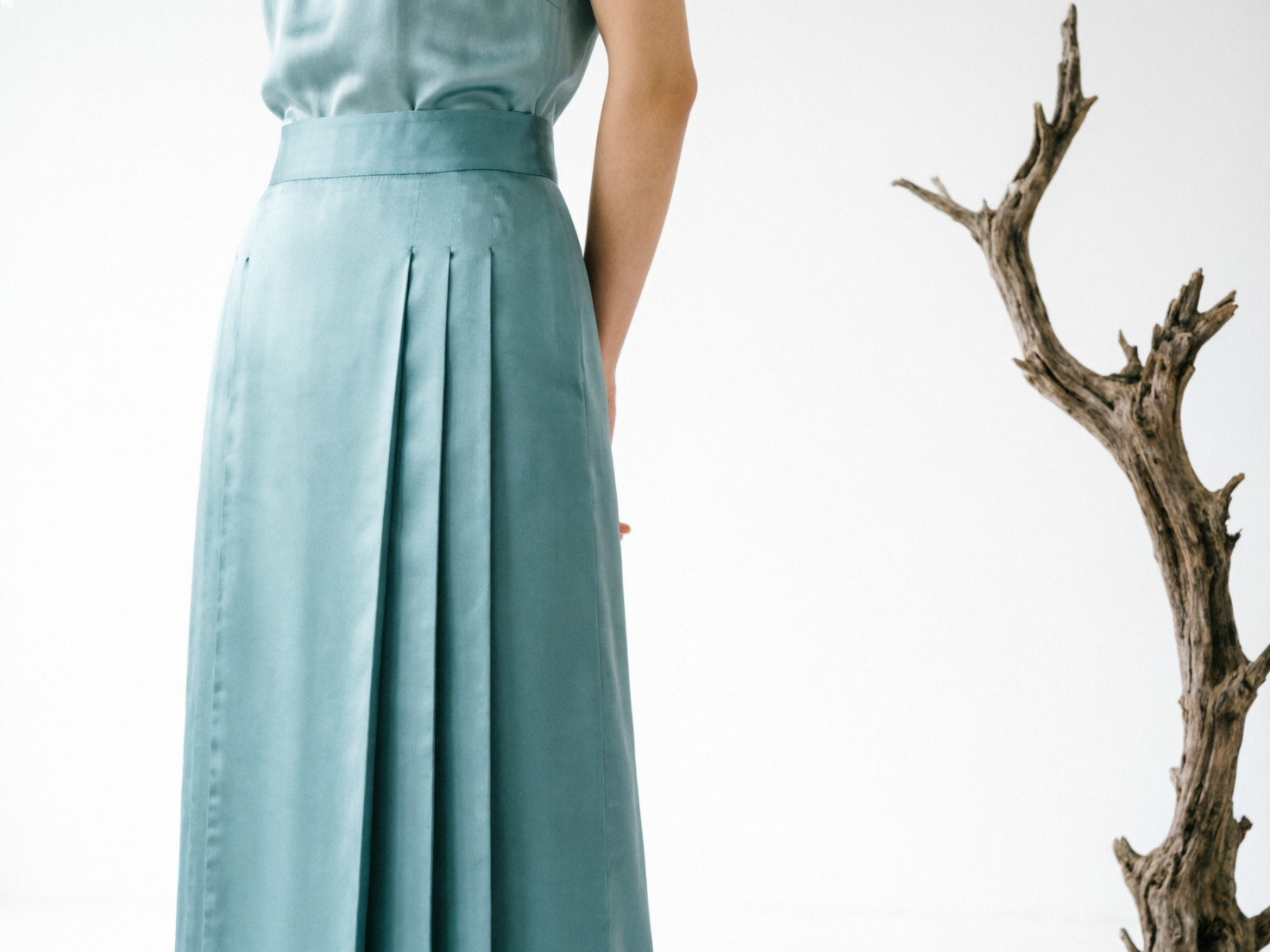 图中展示了一个极简风格的室内场景，背景为纯净的白色。画面左侧是一位背对镜头的女性，她穿着一套淡青色的丝绸质感套装。上衣扎在裙子里，下身是一条带有宽大褶皱的长裙，面料泛着柔和的缎面光泽。画面右侧矗立着一截枯木艺术品，其深褐色的粗糙表皮与女性平滑的衣物形成鲜明对比。构图简洁，留白较多，营造出一种宁静、冷淡的艺术氛围。在 AI 生成的细节方面，存在明显的逻辑缺陷：女性背后的双手部分处理模糊且结构异常，看起来像是手指直接从腰部位置伸出，缺乏完整的手臂连接感；此外，长裙左侧的缝线边缘显得不够自然，与整体光影略有脱节。这些细节揭示了图像的非真实性。