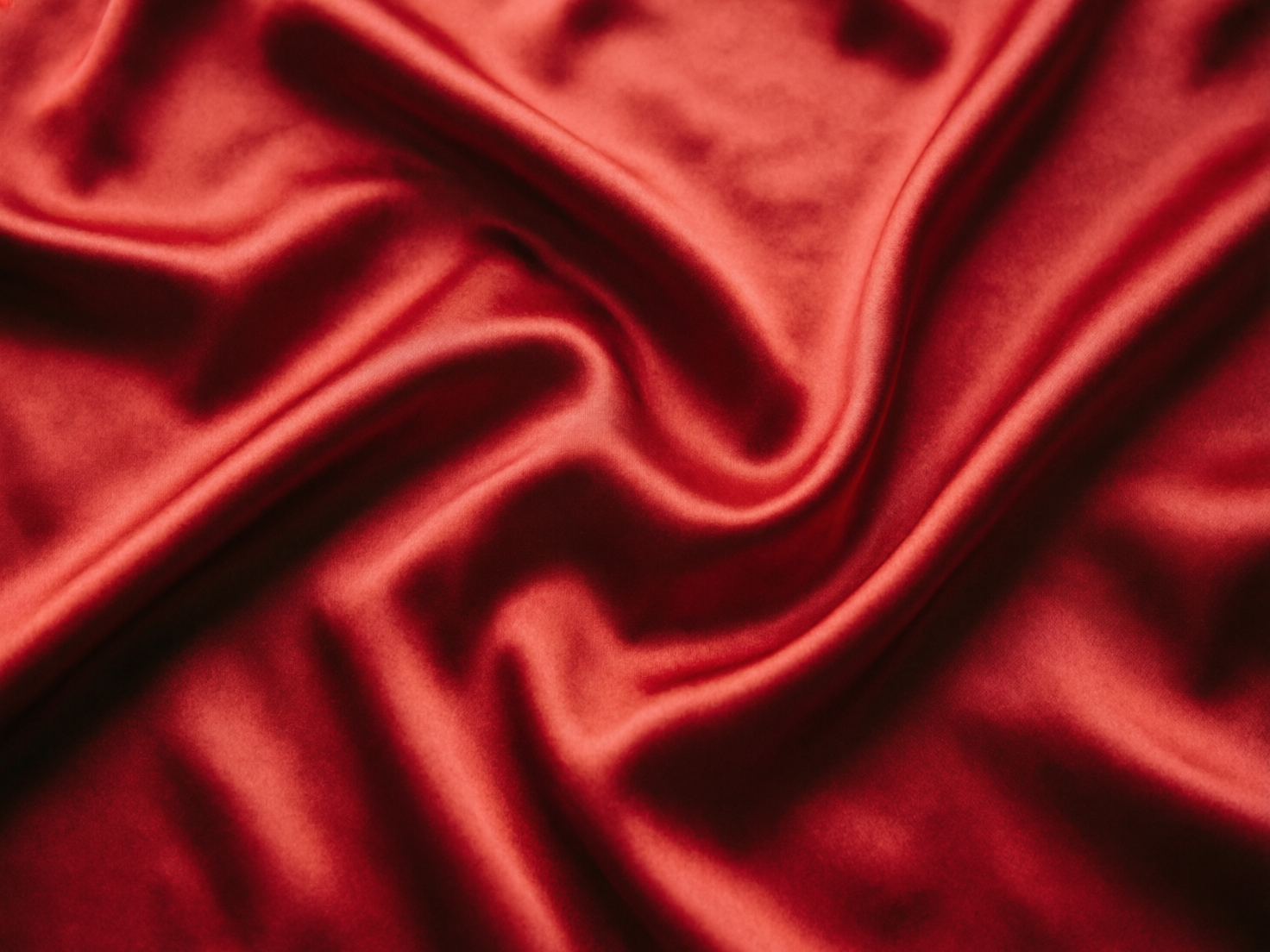 图中展示了一块充满质感的深红色丝绸或缎面织物。整张图片以俯视的角度捕捉了布料自然堆叠、起伏的状态，呈现出如波浪般流畅的曲线和褶皱。光线从斜上方照射，使得布料的凸起部分呈现出明亮的丝滑光泽，而凹陷的褶皱处则隐藏在深沉的阴影中，明暗对比强烈，增强了画面的立体感和流动感。色彩上是统一且浓郁的朱红色，给人一种高贵、温暖且细腻的视觉感受。

在 AI 生成的细节方面，虽然整体质感非常逼真，但仔细观察可以发现一些逻辑上的微小瑕疵：部分褶皱的走向在交汇处显得略微生硬，缺乏真实物理重力下布料层叠的自然垂坠感；此外，画面边缘的某些光影过渡显得过于平滑，略带一种数字渲染的塑料感，缺少了真实纤维织物可能存在的细微纹理或不规则反光。总体而言，这是一张视觉效果极佳的材质模拟图。