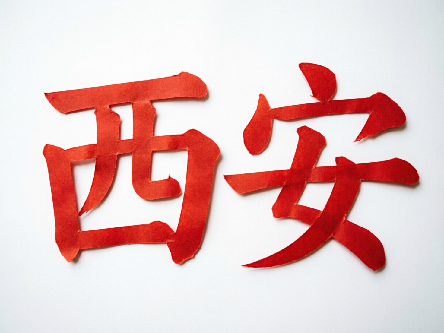 图中展示了在纯白色背景上，用红色纸张剪裁而成的两个汉字“西安”。这两个字占据了画面的中心位置，色彩鲜艳夺目，呈现出一种类似中国传统剪纸或撕纸艺术的质感。字体风格介于楷书和隶书之间，笔画边缘带有不规则的毛边和撕裂感，模拟了手工制作的粗犷效果。

然而，作为一张 AI 生成的图片，画面中存在明显的文字结构缺陷。左侧的“西”字内部结构混乱，原本应为两横的笔画被扭曲成了一个不闭合的钩状，且字形整体向右倾斜。右侧的“安”字问题更为严重，其上方的“宝盖头”完全变形，点和横钩断裂错位，下方的“女”字部首也失去了正常的间架结构，笔画交叉方式极其生硬且不符合汉字书写逻辑，导致文字看起来更像是模仿汉字形状的抽象图形。