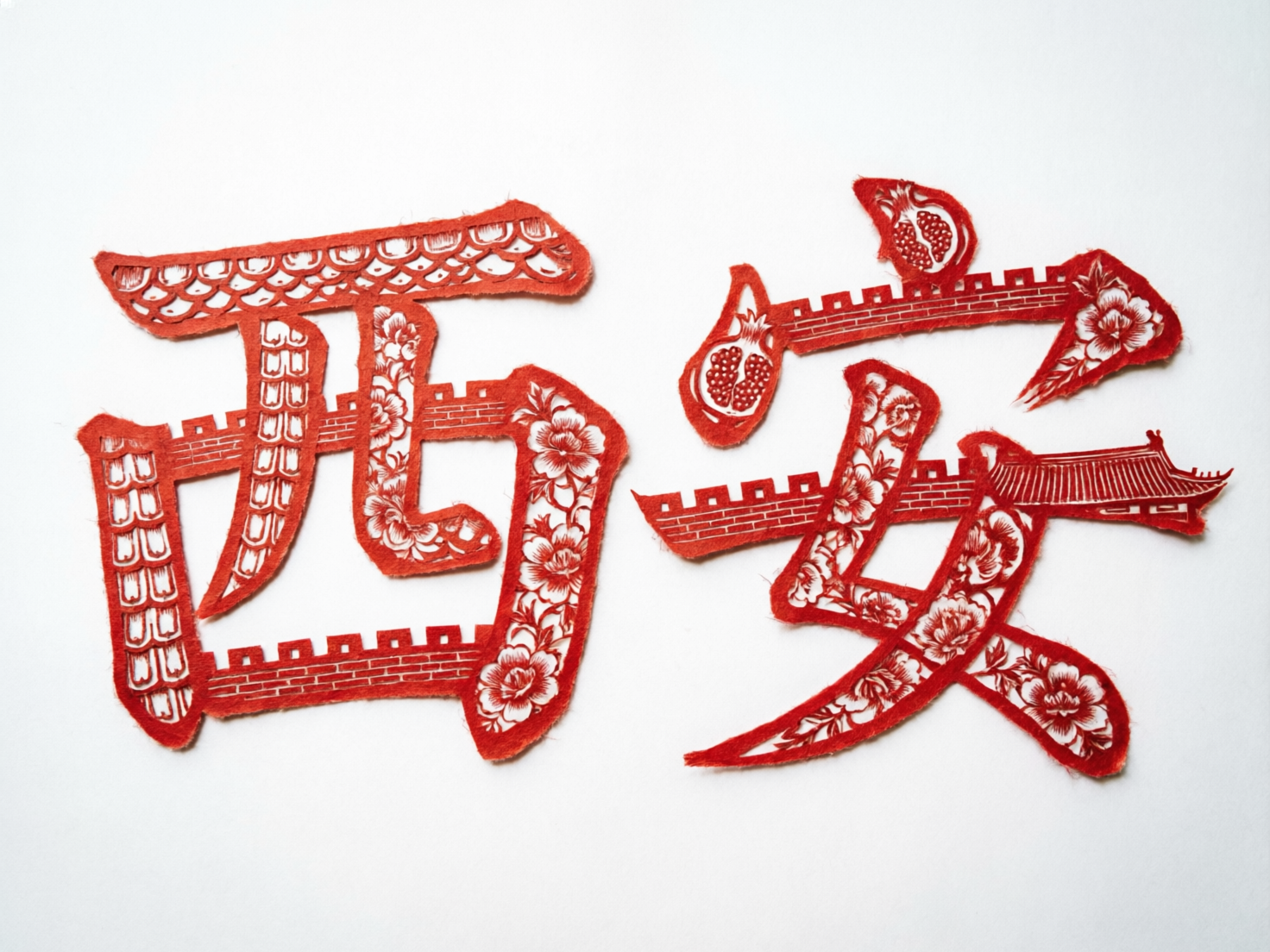 画面中展示了两个具有中国传统剪纸风格的红色艺术字，背景为纯净的白色。这两个字意在表现“西安”二字，笔画内部融合了丰富的文化元素，包括蜿蜒的古城墙、盛开的花卉、石榴图案以及古建筑的屋檐，色彩以正红色为主，边缘带有模拟纸张裁剪的毛边质感。然而，作为 AI 生成的图像，画面存在明显的逻辑与结构缺陷：左侧的“西”字结构扭曲，笔画连接处显得生硬且不符合汉字书写规范；右侧的“安”字则完全解体，笔画断裂并随意堆叠，无法辨认出正确的字体结构。此外，城墙的砖块纹理在转折处出现了透视错误，部分花纹模糊不清，整体呈现出一种形似而神散的视觉效果，未能准确还原真实剪纸的严谨工艺。