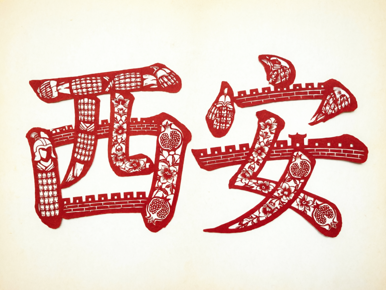 图中展示了一幅模仿中国传统剪纸艺术的红色作品，背景为干净的米白色。画面中心是两个巨大的汉字“西安”，字体风格厚重且富有装饰性。这两个字的笔画内部镂刻了丰富的西安文化元素，包括兵马俑的铠甲纹样、古城墙的垛口、盛开的花卉以及石榴图案。整体色调以大红色为主，白色线条勾勒细节，呈现出浓郁的民俗节日氛围。

然而，作为 AI 生成的图片，画面存在明显的逻辑与结构缺陷。首先，“安”字的结构严重变形，其上方的“宝盖头”断裂成了互不相连的三个碎块，且笔画交接处显得生硬、不符合汉字书写逻辑。其次，笔画中的图案细节较为混乱，部分兵马俑的形象模糊不清，城墙的线条在转折处出现了透视错误和断裂。此外，剪纸边缘的质感略显生硬，缺乏手工剪裁的自然毛边感，部分镂空区域的分布也显得不够均匀。
