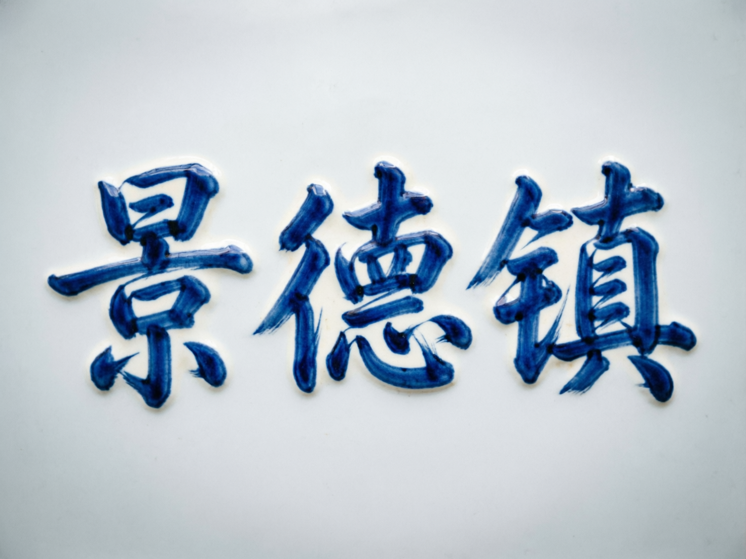 图中展示了在洁白的瓷砖背景上，用青花瓷风格书写的三个汉字“景德镇”。这三个字采用了深蓝色的颜料，笔触模仿了毛笔书法的质感，带有明显的浓淡变化和釉料的立体厚度感，仿佛是刚刚在瓷胎上勾勒完成。构图非常简洁，文字位于画面正中央，四周留有大量的纯白空间。然而，作为 AI 生成的图像，画面中存在明显的文字结构缺陷：第一个“景”字下半部分的笔画发生了扭曲变形，显得杂乱且不符合常规写法；中间的“德”字右半部分结构模糊，缺少了关键的横划和心字底；最后的“镇”字右侧内部的横线数量过多且排列拥挤，失去了汉字应有的间架结构。整体视觉效果虽然传达了陶瓷艺术的韵味，但文字细节上存在明显的逻辑错误。