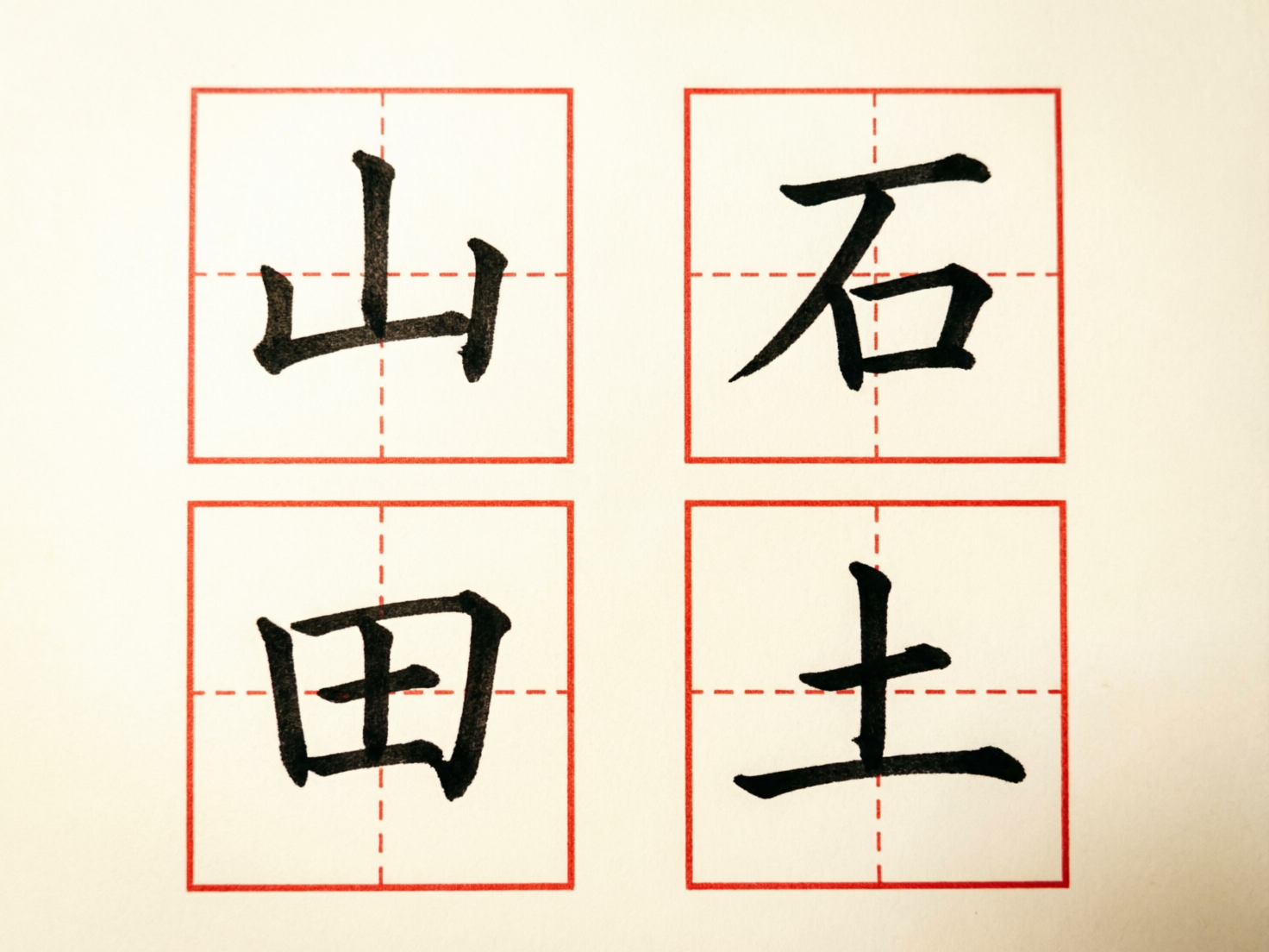 图中展示了一张米黄色背景的习字帖，画面中心排列着四个红色的田字格，每个格子内都有一个黑色毛笔书写的汉字。这四个字从左到右、从上到下依次是“山”、“石”、“田”、“土”。字体呈现楷书风格，笔触带有明显的墨迹质感和粗细变化。构图规整，采用了正面平视的视角，色彩对比鲜明，给人一种传统书法练习的视觉感受。

在 AI 生成的细节方面，存在一些明显的逻辑错误和笔画异常：首先，田字格内部的红色虚线并不连贯，且与外框的连接处显得生硬。其次，汉字的结构存在扭曲，例如“山”字的左侧竖画与横画连接处过于圆滑，缺乏折笔的力度；“田”字内部的横竖交叉点位置偏移，且字形整体向左倾斜；“土”字的第二横末端出现了不自然的墨迹晕染。此外，纸张的纹理在边缘处显得过于平滑，缺乏真实的纸张纤维感。