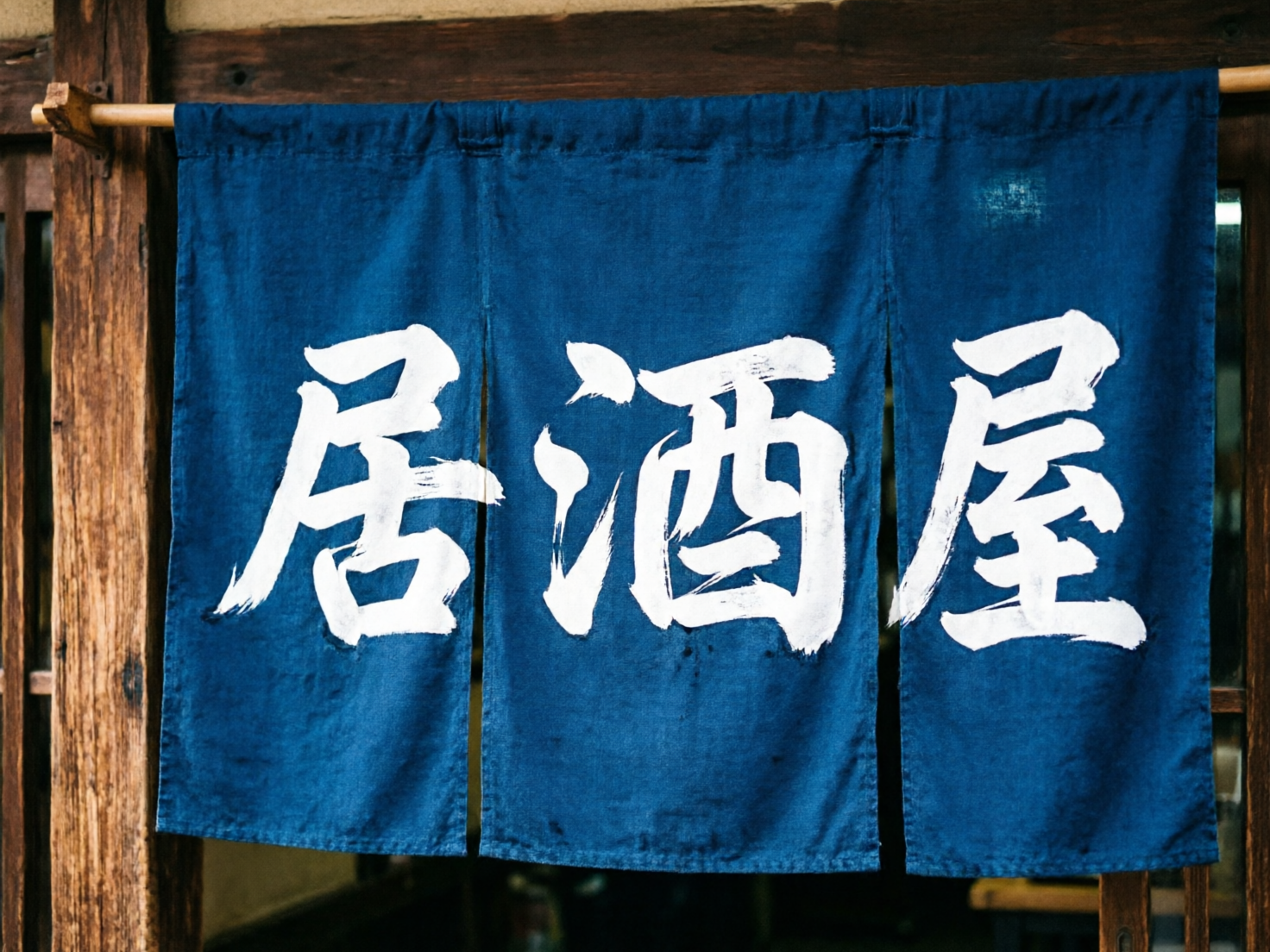 画面中展示了一家日式居酒屋入口处的特写。主体是一块深蓝色的布帘（暖帘），被分成三段，悬挂在粗糙的木质横梁下。布帘上用白色的书法字体写着“居酒屋”三个大字，笔触带有明显的毛刷质感和飞白效果。背景是深褐色的木质门框，隐约可见室内的昏黄灯光和模糊的桌椅轮廓，营造出一种温馨、复古的深夜食堂氛围。色彩对比鲜明，深蓝色与白色的组合非常醒目。

在 AI 生成的细节方面，存在一些明显的逻辑缺陷：首先，中间的“酒”字结构异常，左侧的“氵”旁与右侧部分连接方式生硬，且字形结构不符合常规书法逻辑；其次，布帘上方的悬挂环扣分布不均，左侧的木杆支撑点显得有些悬浮，缺乏真实的受力感；此外，最右侧的“屋”字笔画末端出现了不自然的断裂和模糊，这些都是 AI 在处理复杂汉字结构和物理遮挡关系时常见的瑕疵。
