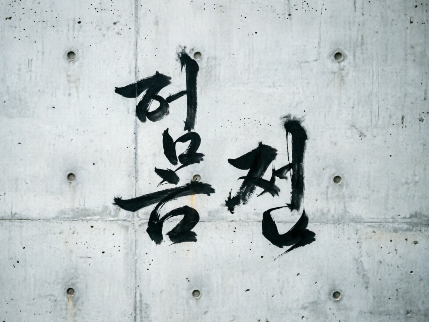 画面中是一面充满工业质感的浅灰色清水混凝土墙面，墙上布满了细小的深色斑点和自然的纹理。墙面被几条横竖交错的细浅接缝分割成规则的矩形块，每个矩形块的角落处都有圆形的凹陷孔洞，呈现出一种现代建筑的冷峻风格。画面中央是用黑色浓墨书写的两个韩文字符，笔触粗犷且富有动感，带有明显的毛笔书法飞白效果，墨迹在粗糙的墙面上显得厚重而有力。然而，作为一张 AI 生成的图片，画面存在明显的逻辑缺陷：黑色的书法字体像是凭空悬浮或直接贴在墙面上的，缺乏与墙面纹理的自然融合感；此外，墙面上的圆形孔洞分布并不完全对称，部分孔洞的边缘模糊，且墙面接缝处的透视关系略显生硬，不符合真实物理环境下的光影逻辑。