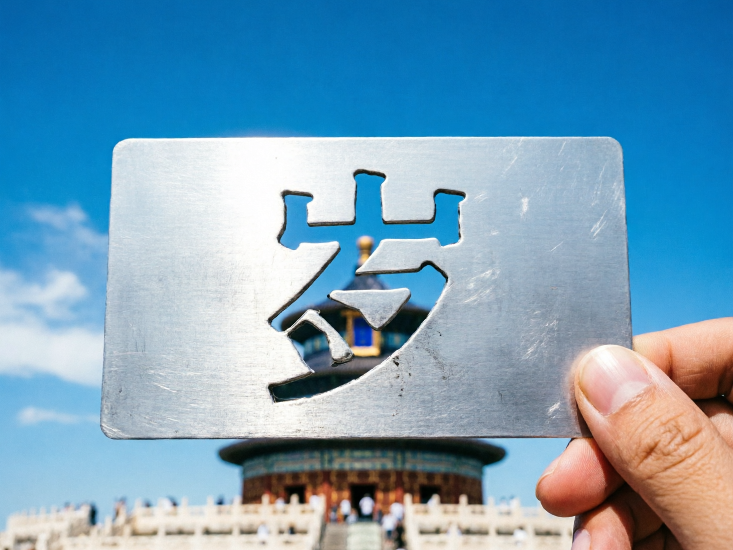 图中展示了一只手拿着一块长方形的金属镂空板，背景是北京天坛的祈年殿。画面构图以金属板为中心，背景的天坛建筑略微虚化。天气晴朗，天空呈现出纯净的深蓝色，点缀着几抹淡淡的白云。金属板表面有细微的划痕和反光，中间镂空出一个汉字形状，通过镂空处可以看到远处的祈年殿金顶。

画面中存在明显的 AI 生成缺陷：首先，金属板中间镂空的汉字并非标准的中文字符，它看起来像是“岁”字或“步”字的变形重组，笔画结构不符合逻辑，且镂空边缘存在不自然的毛刺感。其次，右侧持板的手部细节异常，手指的比例和关节褶皱显得生硬，指甲的形状也略显不自然。最后，背景中天坛下方的游客人群呈现出模糊的色块状，缺乏真实的形体细节，与前景的金属板质感存在透视上的违和感。