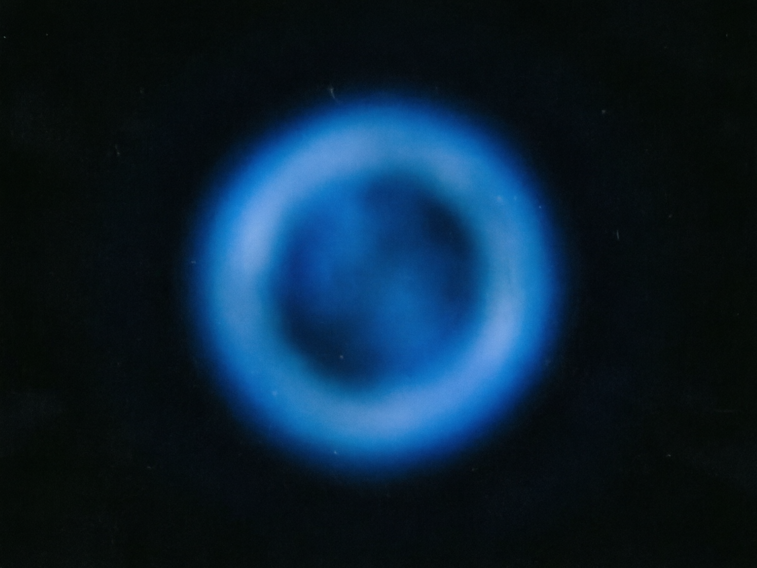 图中展示了一个位于纯黑色背景中央的蓝色发光圆环，呈现出一种深邃且神秘的宇宙感。这个圆环并非线条清晰的实心物体，而是由柔和的蓝色光晕组成，看起来像是一团发光的星云或某种能量场。圆环的中心区域颜色较深，呈现出一种幽暗的深蓝色，而外圈则散发出明亮的浅蓝色光芒，边缘带有明显的模糊感和扩散效果。

从 AI 生成的角度来看，画面整体构图极其简单，缺乏精细的纹理细节。圆环的形状虽然大致对称，但光影分布略显生硬，缺乏自然天体应有的层次感和随机性。画面中存在一些细微的白色噪点和杂质，这可能是模拟胶片质感时产生的瑕疵，也可能是生成算法在处理纯色背景时的不稳定性表现。整体视觉效果更像是一个简单的图形滤镜产物，而非真实拍摄的物理现象。
