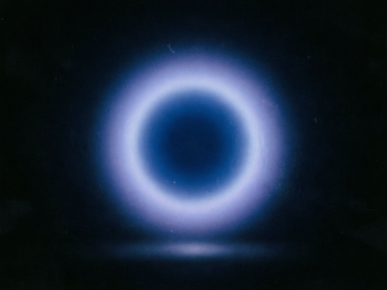 图中展现了一个充满科幻感与神秘气息的视觉场景。画面的主体是一个悬浮在深邃黑色背景中央的巨大发光圆环。这个圆环呈现出柔和的渐变色调，从中心深邃的暗蓝色向外扩散，逐渐转变为明亮的浅蓝色和淡紫色荧光。圆环的边缘带有明显的晕染效果，仿佛光芒在空气中微微震荡。在圆环下方的地面上，可以看到一抹淡淡的蓝色倒影，增强了空间的立体感。

从 AI 生成的角度观察，画面整体风格偏向极简抽象，但也存在一些细微的生成痕迹。首先，背景的黑色并不纯净，散布着一些类似胶片噪点或灰尘的白色细小杂质，这可能是 AI 模拟复古质感时产生的随机像素。其次，发光圆环的圆周并非绝对完美的几何圆，在边缘处存在轻微的厚薄不均和光影断层。此外，底部的倒影与上方圆环的亮度比例略显失调，透视关系上显得有些生硬，缺乏真实物理环境下的自然过渡。