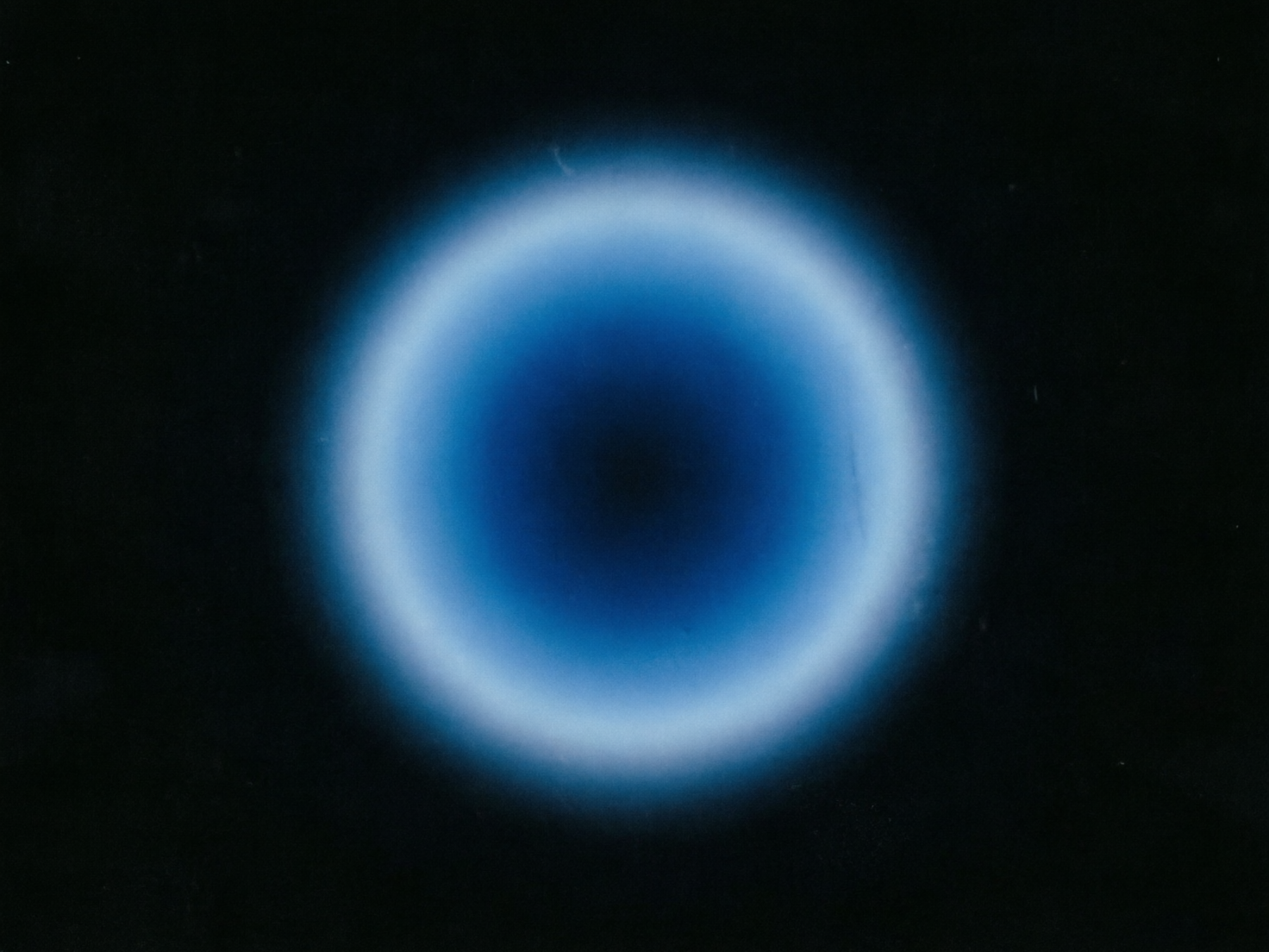 图中展示了一个位于纯黑色背景中央的圆形发光体，呈现出一种深邃而神秘的极简主义风格。这个圆形物体由内向外呈现出明显的色彩梯度变化：最中心是一个深邃的黑色圆心，向外逐渐过渡为浓郁的深蓝色，再演变为明亮的浅蓝色，最外圈则是一层柔和的白色光晕，整体看起来像是一个发光的瞳孔或是一个遥远的星云。构图非常对称，光影过渡细腻且具有朦胧的磨砂质感。在 AI 生成的细节方面，虽然整体视觉效果较好，但可以观察到光圈的边缘并非完美的正圆，存在轻微的形状扭曲和不规则的毛边。此外，黑色背景中分布着一些细微的白色杂质点和类似划痕的纹理，这可能是 AI 在模拟胶片质感时产生的随机噪点，而非真实的物理痕迹。