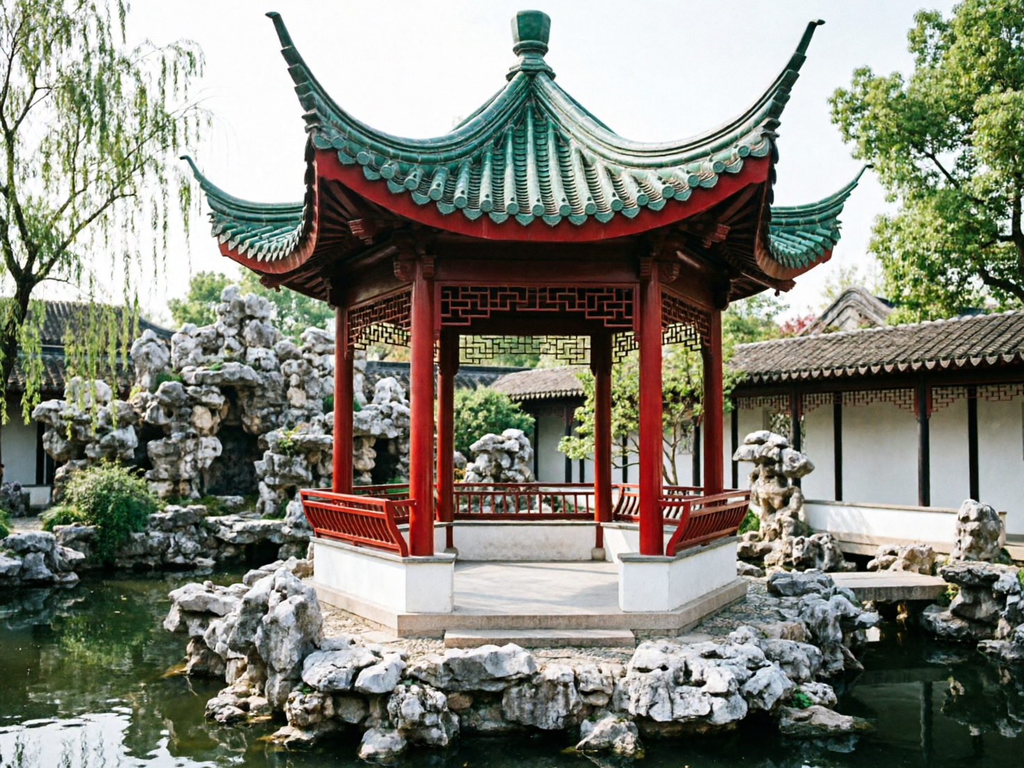 图中展示了一座位于中式园林湖泊中央的古典凉亭。凉亭采用典型的飞檐翘角设计，屋顶覆盖着青绿色的瓦片，边缘衬以红色装饰，亭身由几根粗壮的红色木柱支撑。凉亭坐落在由嶙峋怪石堆砌而成的基座上，四周环绕着平静的池水，背景可见垂柳、假山和白墙黑瓦的连廊，整体色调清新自然，充满古朴韵味。

然而，画面中存在多处明显的 AI 生成缺陷：首先，凉亭的结构逻辑混乱，屋顶的瓦片排列在边缘处显得模糊且不连贯，左侧翘角的弧度与右侧不对称。其次，凉亭内部的红色护栏与柱子的连接处透视异常，线条生硬且不符合物理结构。最明显的错误在于凉亭下方的石块基座，部分石头呈现出液化般的模糊感，且右侧通往连廊的小桥与岸边的衔接处完全断裂，悬浮在水面上，缺乏合理的支撑结构。