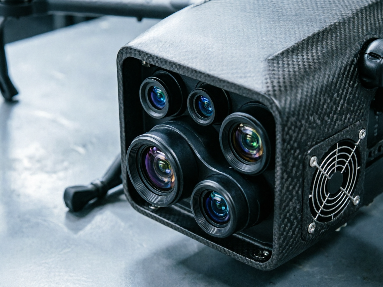 图中展示了一个具有工业科技感的黑色设备近景，看起来像是一台配备了多镜头系统的无人机载荷或专业摄像机。设备外壳采用了明显的碳纤维纹理，呈现出深灰色和黑色的交织感，侧面安装有一个带有金属保护网的小型散热风扇。镜头组位于一个矩形框架内，包含五个大小不一的圆形镜头，镜片反射出蓝色、紫色和绿色的幽光。背景是模糊的浅灰色平滑表面，左侧隐约可见类似无人机起落架的支架结构。画面整体色调偏冷，光影对比鲜明。在 AI 生成的细节方面，存在明显的逻辑缺陷：镜头组的排列显得杂乱且缺乏光学对称性，部分镜头的边缘与底座融合得不够自然；右侧散热风扇的金属网罩线条略显扭曲，且固定螺丝的位置分布不均，体现了 AI 在处理复杂机械结构时的不严谨。