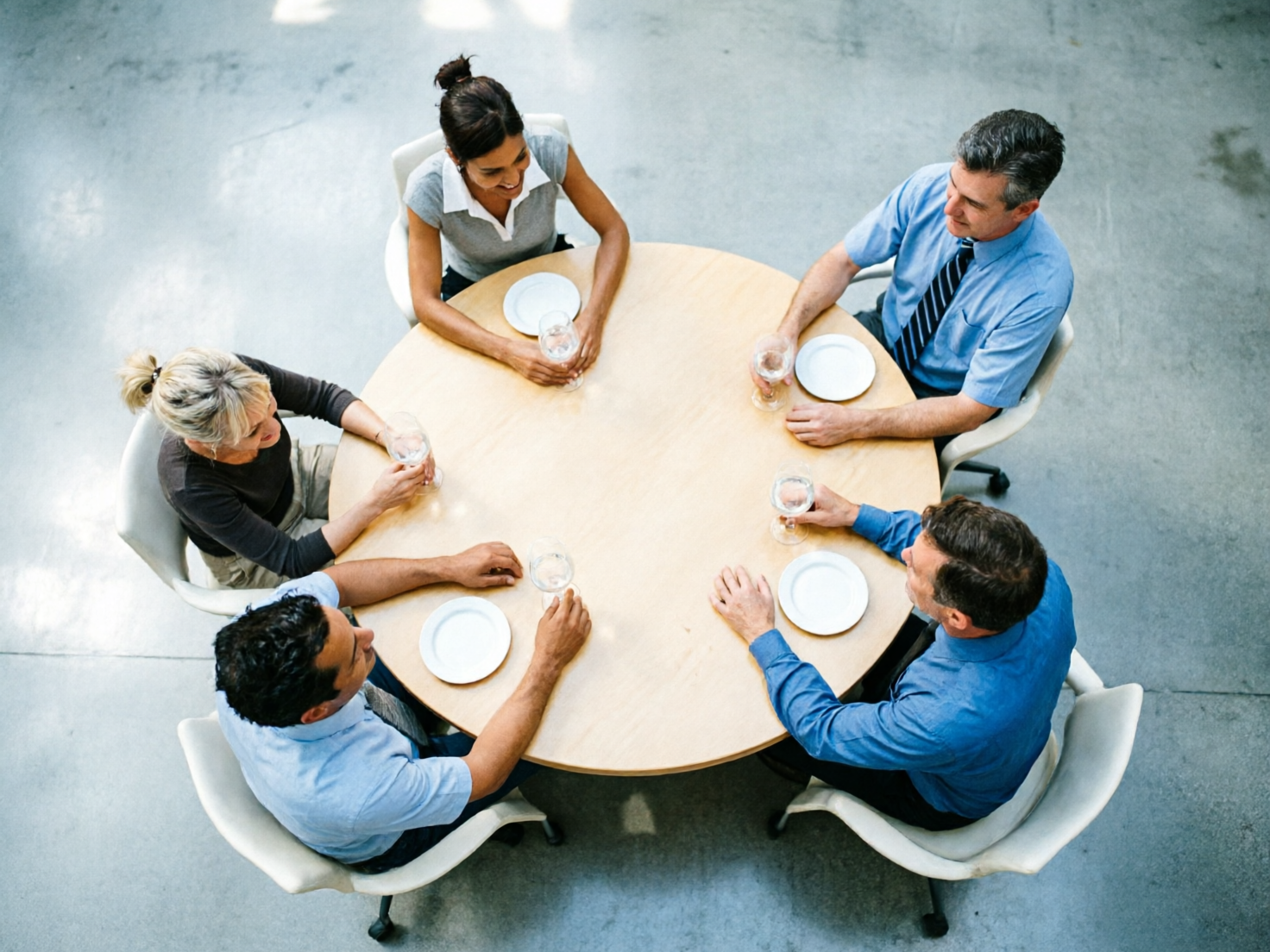 图中展示了一个从高处俯拍的室内场景，五名穿着商务休闲装的男女围坐在一张浅色木质圆桌旁，似乎正在进行商务午餐或会议。背景是灰色的水泥地面，整体色调明亮且偏冷。画面中包含三名男性和两名女性，他们身穿蓝色衬衫、灰色上衣等职业服装，每人面前都摆放着一个白色空盘子和一杯水。

然而，这张图片存在多处明显的 AI 生成缺陷。首先，人物的手部结构严重畸形，例如右下方男性的右手手指数量异常且扭曲，左上方女性握杯子的手部线条模糊。其次，桌上的餐具分布逻辑混乱，盘子和水杯的透视关系不自然。最严重的缺陷在于人物的身体连接：左下方男性的左臂似乎直接消失在桌子边缘，而右上方男性的身体比例与座椅的衔接显得非常生硬。此外，部分人物的面部五官模糊不清，缺乏真实的纹理细节。