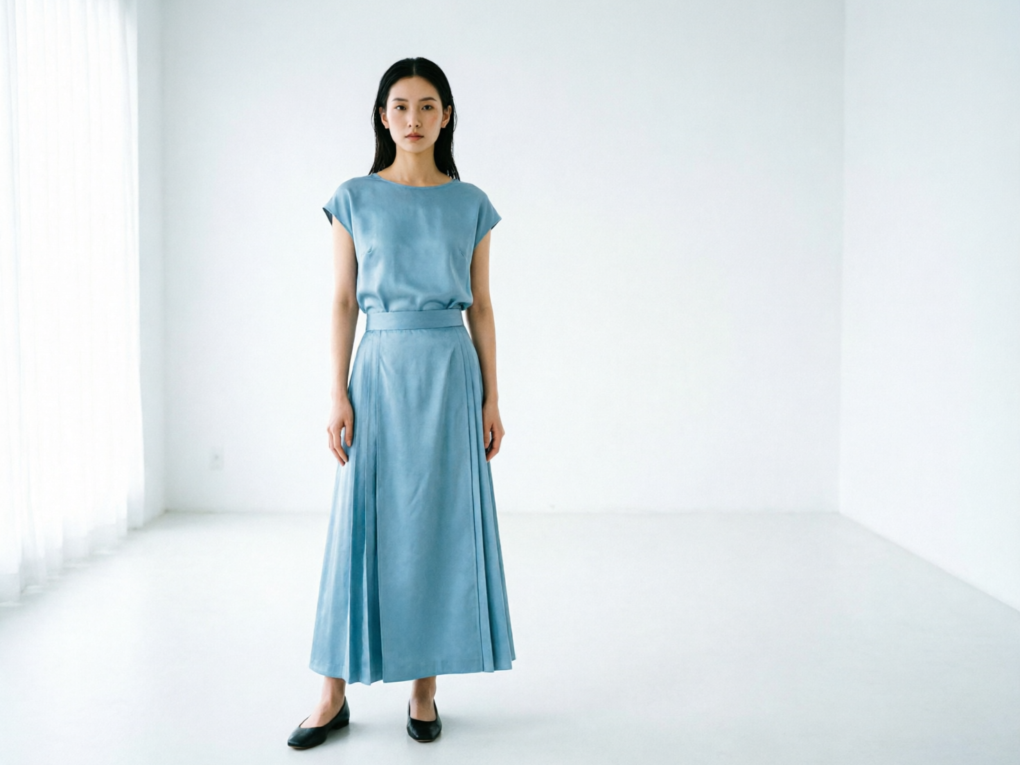 图中展示了一位留着黑色长直发的亚洲女性，她正站在一个极简主义风格的纯白房间中央。她身穿一套灰蓝色的绸面套装，上衣是短袖圆领小衫，下身是带有褶皱设计的长裙，脚上穿着一双黑色平底皮鞋。光线从画面左侧的白色纱帘窗户透入，营造出一种清冷、静谧的氛围。构图简洁，人物位于画面中心，背景是空旷的白墙和地板。在 AI 生成的细节方面，存在明显的逻辑缺陷：女性的双手垂在身体两侧，但手指的结构模糊且扭曲，呈现出不自然的粘连感；此外，她右脚的鞋子形状略显畸形，与脚部的连接处透视关系不够自然。尽管整体色调和谐，但这些肢体细节揭示了其人工智能生成的痕迹。