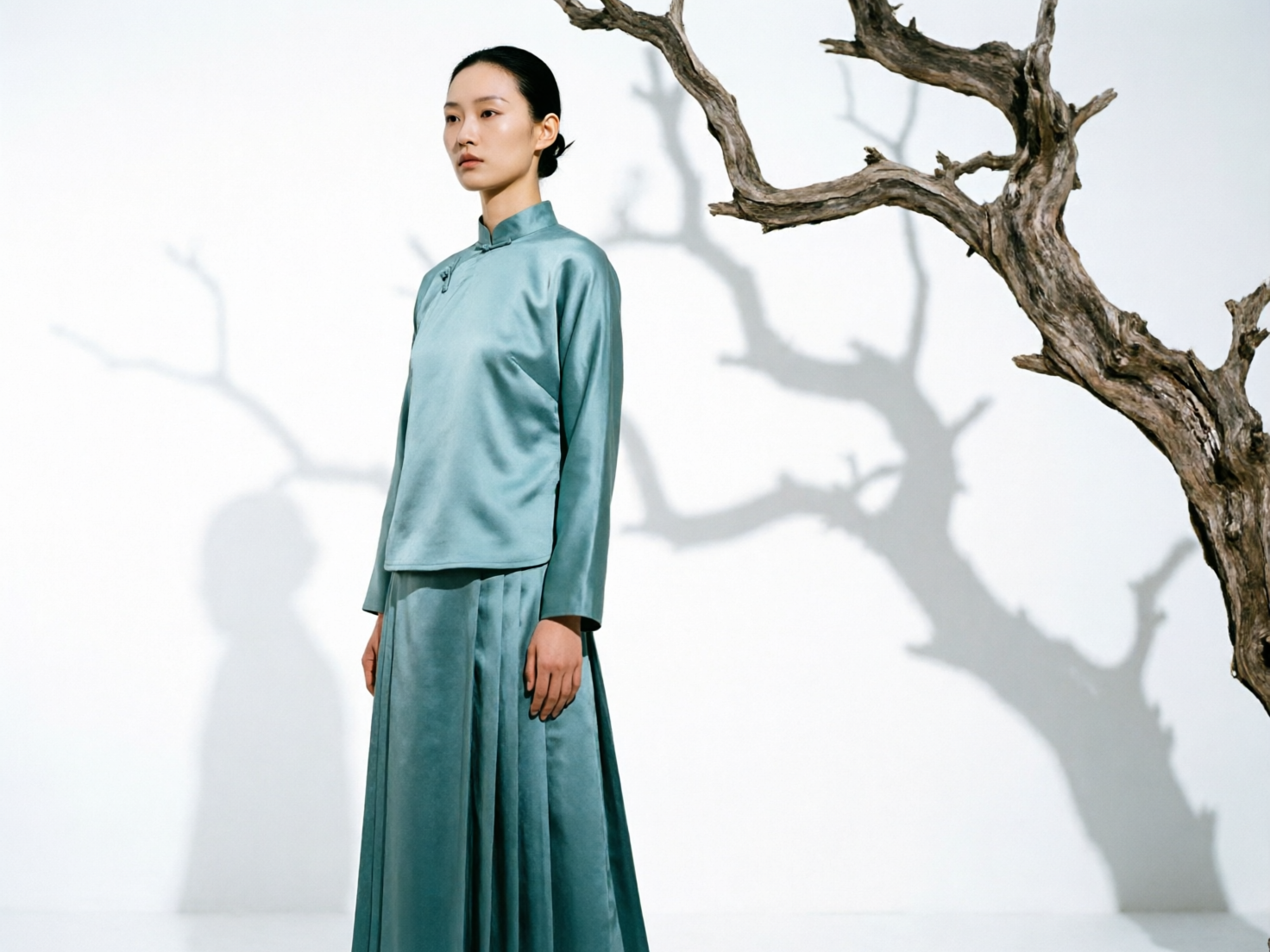 图中展现了一位具有东方韵味的女性模特，她侧身站立在纯白色的背景前。模特留着整洁的黑色盘发，身穿一套青色绸缎质感的改良旗袍上衣和同色系的长裙，面部表情清冷且疏离，目光投向画面左侧。画面右侧是一株枯木，其苍劲的枝干向中心延伸，在白色背景墙上投射出斑驳且巨大的阴影，营造出一种极简而富有禅意的艺术氛围。整体色调以冷青色和白色为主，视觉效果干净利落。

在 AI 生成的细节方面，存在一些明显的逻辑缺陷：首先，画面左侧墙上的影子与右侧实物枯木的形态并不完全对应，且影子中出现了一个模糊的人头轮廓，但其位置与模特本体的站位不符。其次，模特的左手（画面右侧）手指细节略显生硬，指缝处理不够自然。此外，枯木的枝干在接近画面边缘处有不自然的断裂感，背景的地面与墙面交界处过于模糊，缺乏真实的透视空间感。