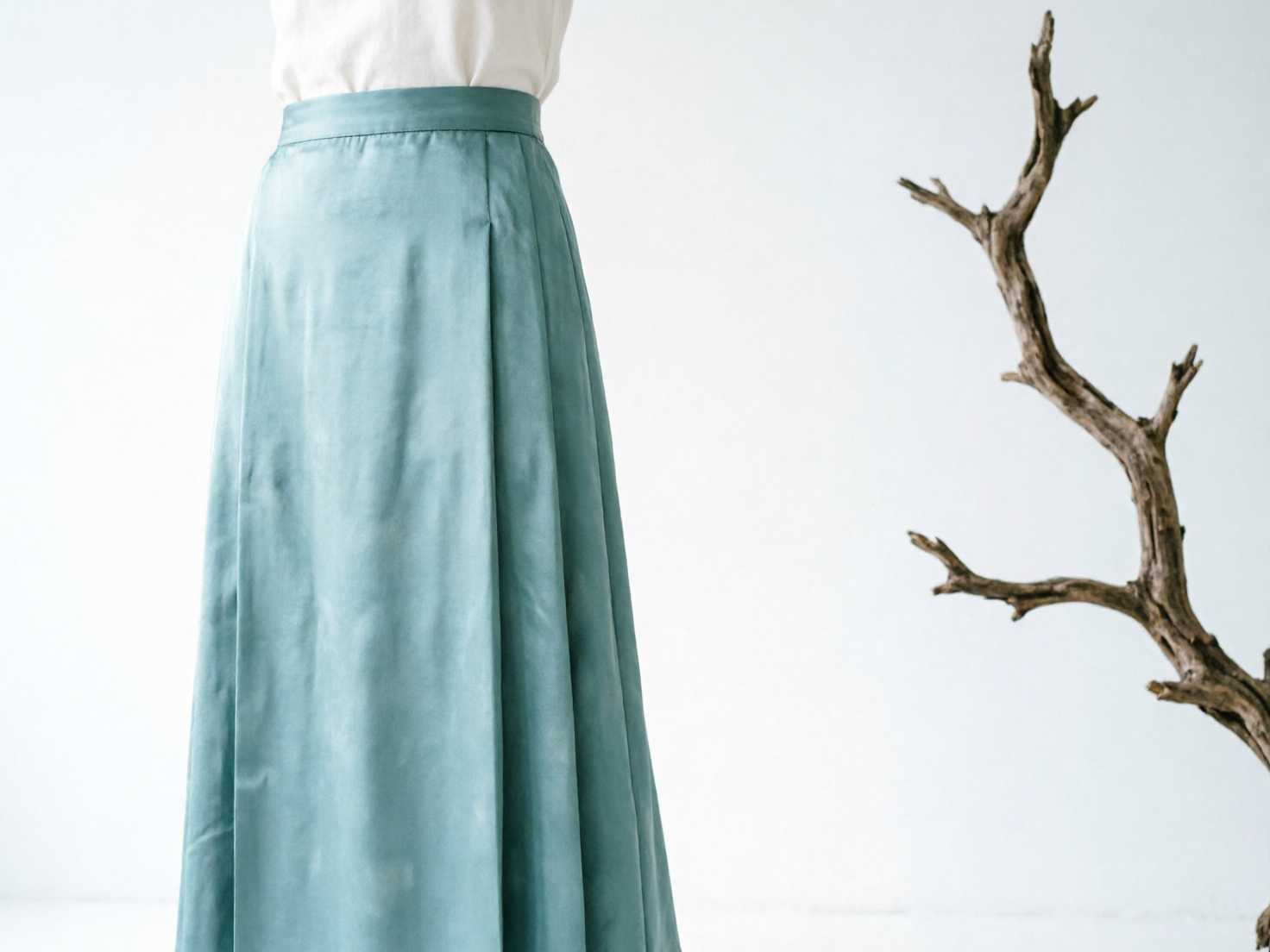 图中展示了一位女性的下半身着装，背景是纯净的白色，整体呈现出简约、冷淡的艺术风格。画面左侧占据主体的是一条长及脚踝的青蓝色绸缎半身裙，裙子带有细腻的光泽感，腰部剪裁合体，裙摆自然下垂并带有几处工整的褶皱。女性上身穿着一件米白色的无袖或短袖上衣，并将其扎入裙腰中。画面右侧是一株形态扭曲、干枯的褐色枯木枝干，作为装饰元素从边缘延伸进来，与柔软的衣物材质形成鲜明对比。

在 AI 生成的细节方面，存在一些明显的逻辑缺陷：首先，裙子的布料纹理分布不均，表面出现了类似水渍或褪色的不规则斑块，这在真实的丝绸面料中并不常见。其次，裙摆左侧的边缘线条显得过于生硬且缺乏自然的垂坠弧度，看起来像是一块平整的板材。此外，右侧的枯木枝干在空间透视上略显突兀，与背景的衔接处缺乏自然的阴影过渡，使其看起来像是后期粘贴上去的素材。