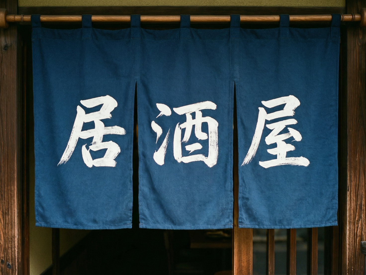 图中展示了一家传统风格日本居酒屋的入口。画面中心是一幅深蓝色的布帘（暖帘），悬挂在横贯画面上方的木质横杆上。布帘分为三片，每片上各有一个白色的手写书法体汉字，从左至右合起来是“居酒屋”三个字。布帘的质感粗糙，带有明显的织物纹理，白色字迹呈现出毛笔刷过的枯笔效果。背景是深色的木质门框，隐约可以看到门帘后方昏暗的室内环境。

在 AI 生成细节方面，画面整体观感较为自然，但存在一些逻辑瑕疵：最左侧和最右侧的布帘挂环与木杆的连接处显得模糊，缺乏真实的物理受力感。此外，右侧木质门框的边缘出现了一些不自然的重影和纹路断裂，横杆最右端的固定件结构模糊，不符合现实中的木工逻辑。文字虽然清晰可辨，但“酒”字的偏旁部首比例略显失调，笔画末端的细节处理带有典型的 AI 算法平滑痕迹。