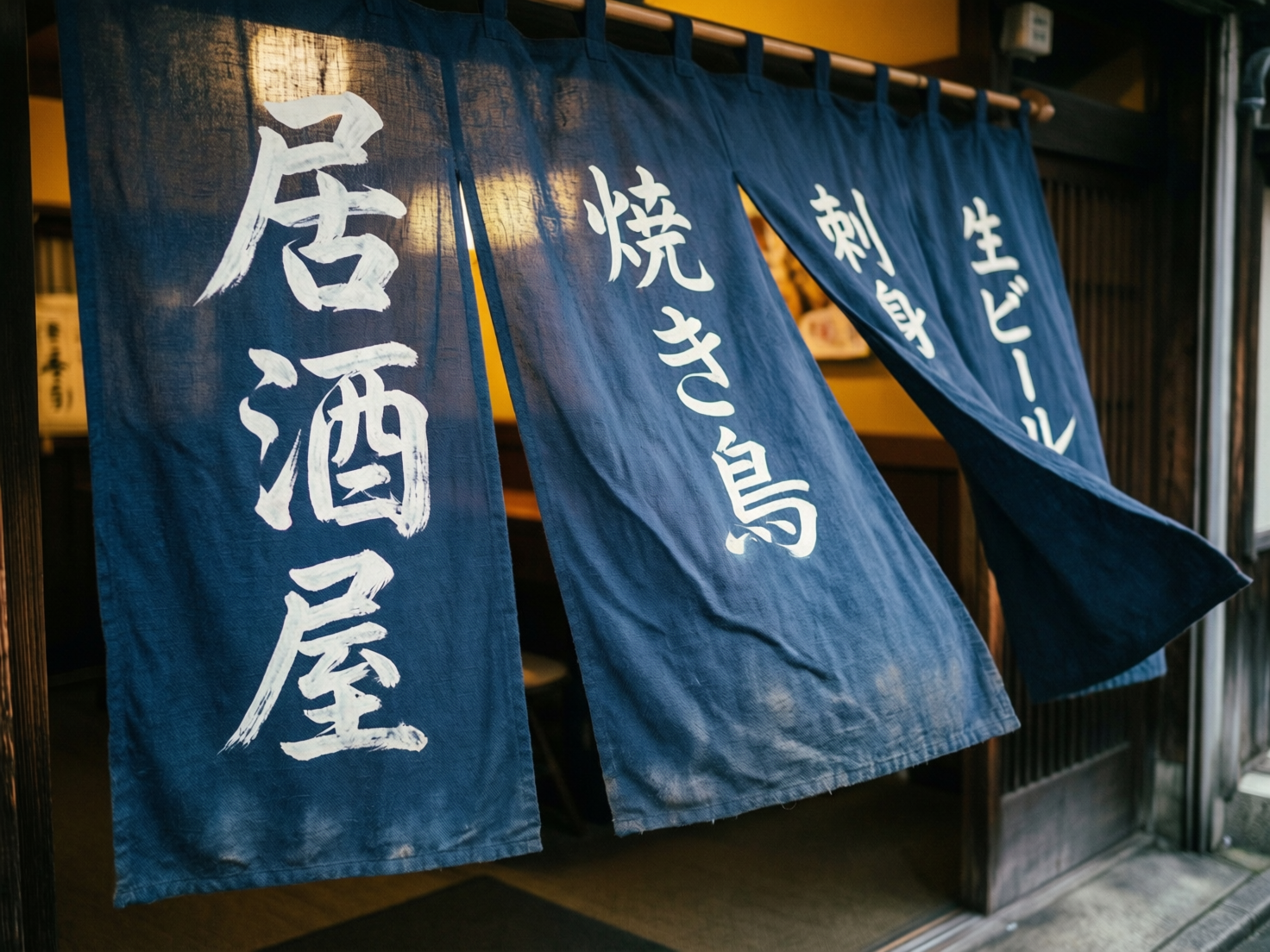 图中展示了一家日式居酒屋入口处的近景。画面主体是悬挂在木质横杆上的深蓝色布帘（暖帘），布帘由四块长方形织物组成，正随风轻轻飘动。布帘上用白色手写体书法印有日文字样，从左至右分别写着“居酒屋”、“焼き鳥”（烤鸡肉串）、“刺身”和“生ビール”（生啤酒）。背景透出店内温暖的黄色灯光和模糊的木质内装，营造出一种深夜食堂的温馨氛围。构图采用斜向视角，焦距集中在最左侧的布帘上，背景带有自然的虚化效果。在 AI 生成的细节方面，存在明显的文字和结构缺陷：最左侧“居酒屋”的“屋”字下半部分笔画杂乱且不完整；中间“焼き鳥”的“鳥”字底部横画缺失，结构扭曲；此外，布帘上方的吊环与横杆的穿插关系在透视上略显生硬，部分布料的褶皱走向与风吹的方向不完全吻合。