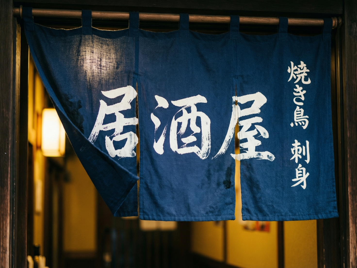 图中展示了一家日式居酒屋入口处的特写。画面主体是一块深蓝色的布帘（暖帘），悬挂在木质横杆上。布帘由三片组成，最左侧的一片微微掀起，露出后方透着暖黄色灯光的室内环境。布帘上印有醒目的白色手写体汉字“居酒屋”，右侧则竖排印着“焼き鳥 刺身”（烤鸡肉串 刺身）字样。整体色调以深蓝、木质棕和暖橘色为主，营造出一种温馨、深夜食堂般的氛围。在 AI 生成的细节方面，存在明显的文字逻辑错误：右侧竖排文字中的“鳥”字下半部分笔画杂乱，并非标准字体；“刺身”二字也存在笔画粘连和结构扭曲。此外，布帘左侧掀起的褶皱边缘过于平滑，缺乏自然的织物重力感，且布帘上方的吊环与横杆的穿插关系在透视上略显生硬。