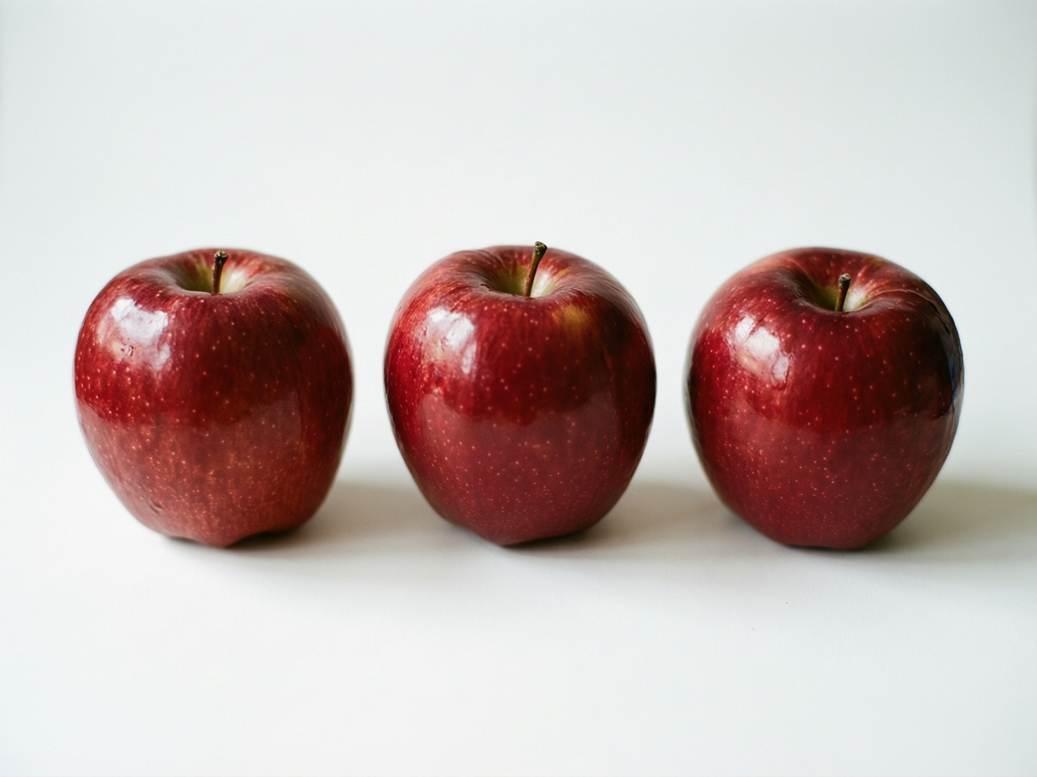 图中展示了三个深红色的苹果，它们在纯白色的背景上横向整齐排列。这些苹果表面带有光泽，仿佛刚被清洗过，反射出柔和的白光，果皮上布满了细小的浅色斑点。构图简洁，光线从上方照射，在苹果下方投射出淡淡的阴影，营造出一种极简主义的静物感。然而，画面中存在明显的 AI 生成缺陷：最左侧苹果的底部轮廓显得有些扭曲，与平面的接触感不够自然；中间苹果的果梗连接处显得生硬，缺乏真实的纹理过渡；最右侧苹果的形状过于圆润对称，且其下方的阴影边缘出现了不自然的模糊和断层。整体而言，虽然色彩浓郁诱人，但物体与背景的融合处略显生硬，缺乏真实摄影中的物理质感。