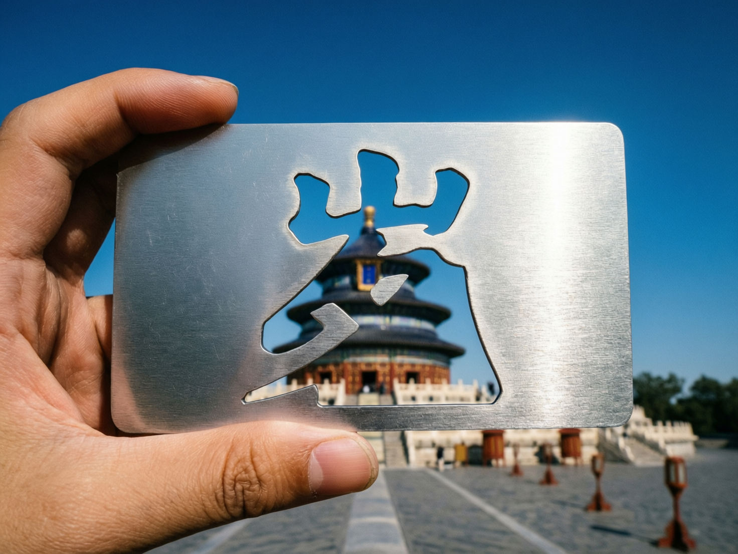 图中展示了一只左手拿着一张银色金属镂空卡片，背景是北京天坛的祈年殿。画面构图以手持卡片为视觉中心，卡片中央镂空出一个类似汉字“岁”或“岸”的变形符号，透过镂空部分可以看到远处被模糊处理的祈年殿。背景天空呈现纯净的深蓝色，地面是灰色的石板路。在 AI 生成的细节方面，存在明显的逻辑缺陷：首先，卡片镂空部分的中间有一个悬空的金属块，在现实中由于没有支撑点，它不可能悬浮在空中；其次，持卡人的手指纹路略显模糊且比例不够自然；此外，镂空的文字形状并不规范，难以辨认具体的汉字。整体色调明亮，光影效果集中在金属卡片的拉丝质感上。