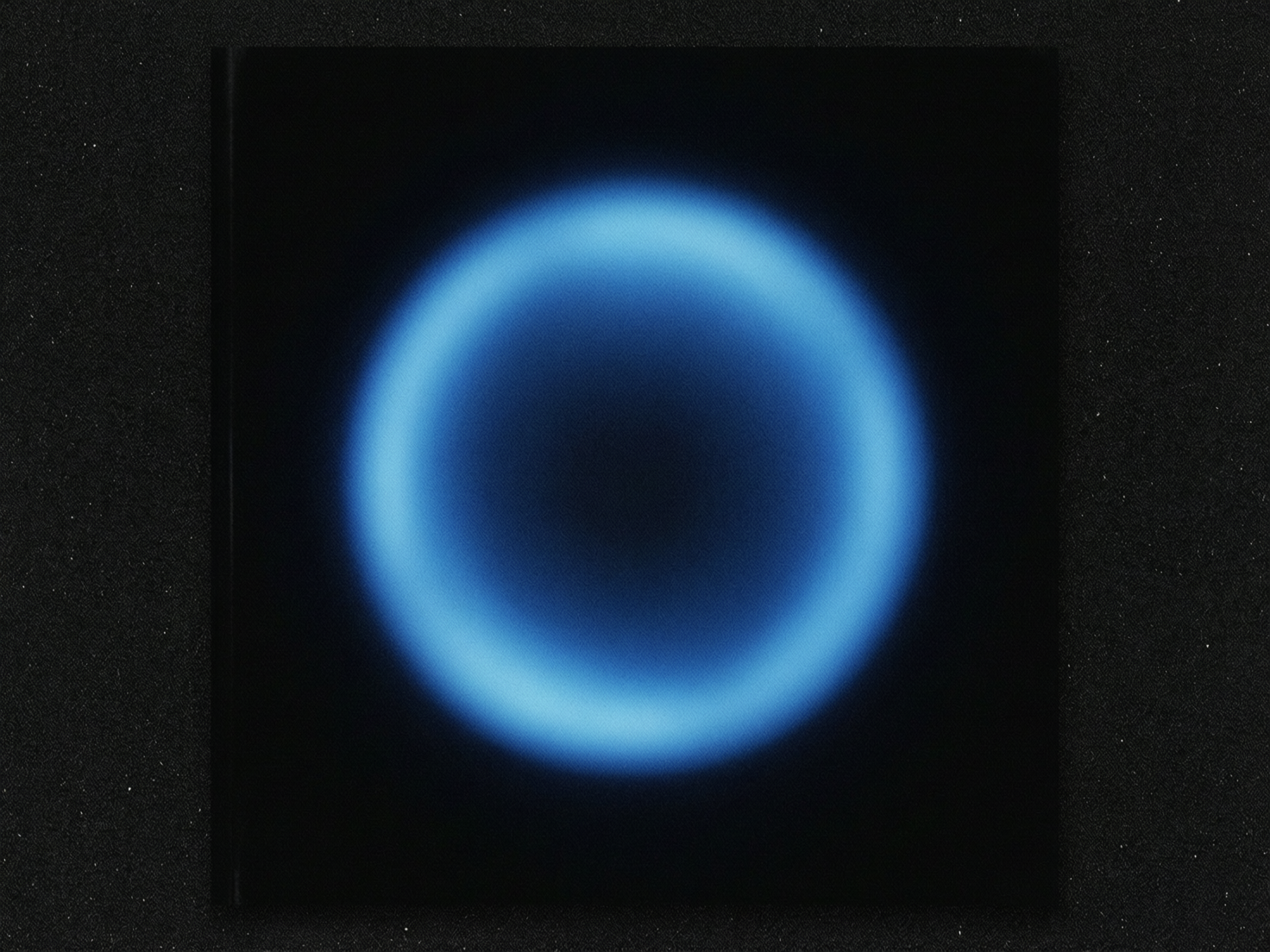 图中展示了一个极简主义风格的抽象画面。正中心是一个明亮的蓝色发光圆环，悬浮在深邃的黑色背景之上。这个圆环呈现出一种柔和的霓虹感，边缘带有明显的模糊和晕染效果，中心则是空洞的黑色，整体像是一个发光的星环或某种能量场。画面的构图非常对称，视觉重心完全集中在中央的蓝色光圈上。背景并非纯黑，而是带有细腻的噪点和颗粒感，模拟了复古胶片或磨砂纸张的质感。在黑色背景的边缘，隐约可以看到类似书籍装订的压痕，暗示这可能是一张唱片封面或画册的设计图。作为 AI 生成的图像，该图在几何对称性上表现较好，但仔细观察可以发现，蓝色圆环的内径边缘光影分布略显不均，左侧的光晕比右侧稍厚，这种细微的非绝对对称是 AI 在处理模糊光影时常见的特征。此外，背景中的噪点分布在极边缘处略显生硬，缺乏自然的物理渐变。